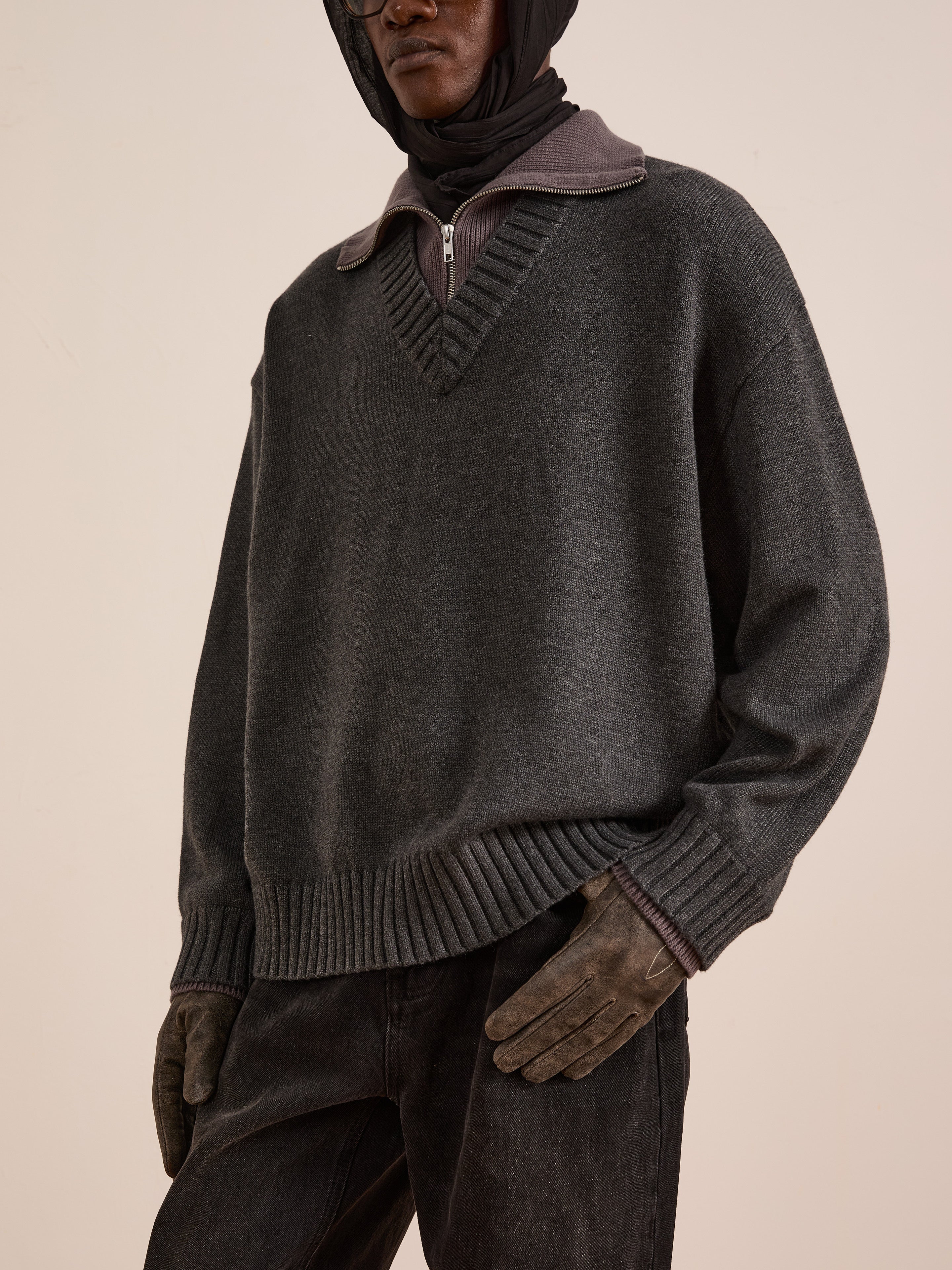 A person showcases practical cold-weather layering in the FOUND Double Layer Sweater—a dark grey wool blend—over a zip-collared shirt, paired with dark jeans, brown gloves, and a hooded head covering, standing against a plain background.