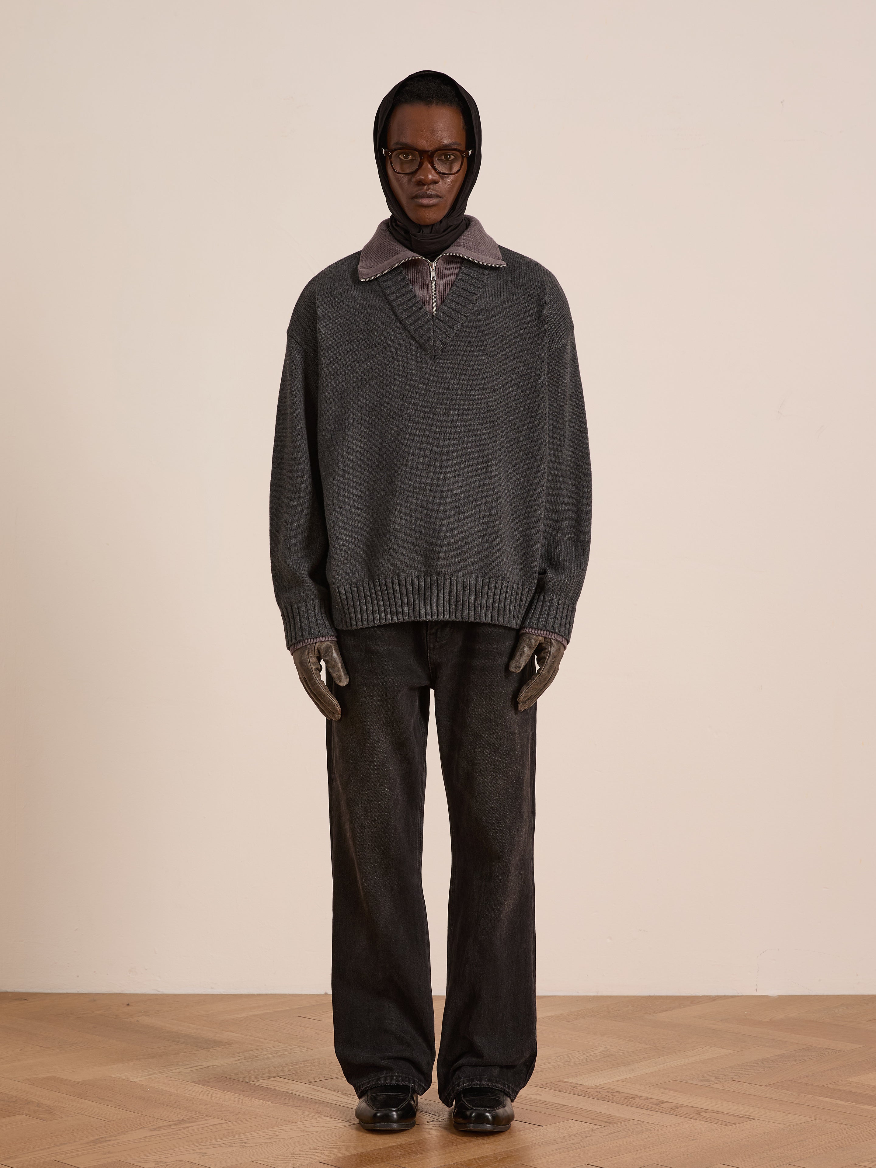 A person stands indoors by a light wall and wooden floor, wearing the FOUND Double Layer Sweater over a collared shirt with dark pants, gloves, glasses, and a hood.