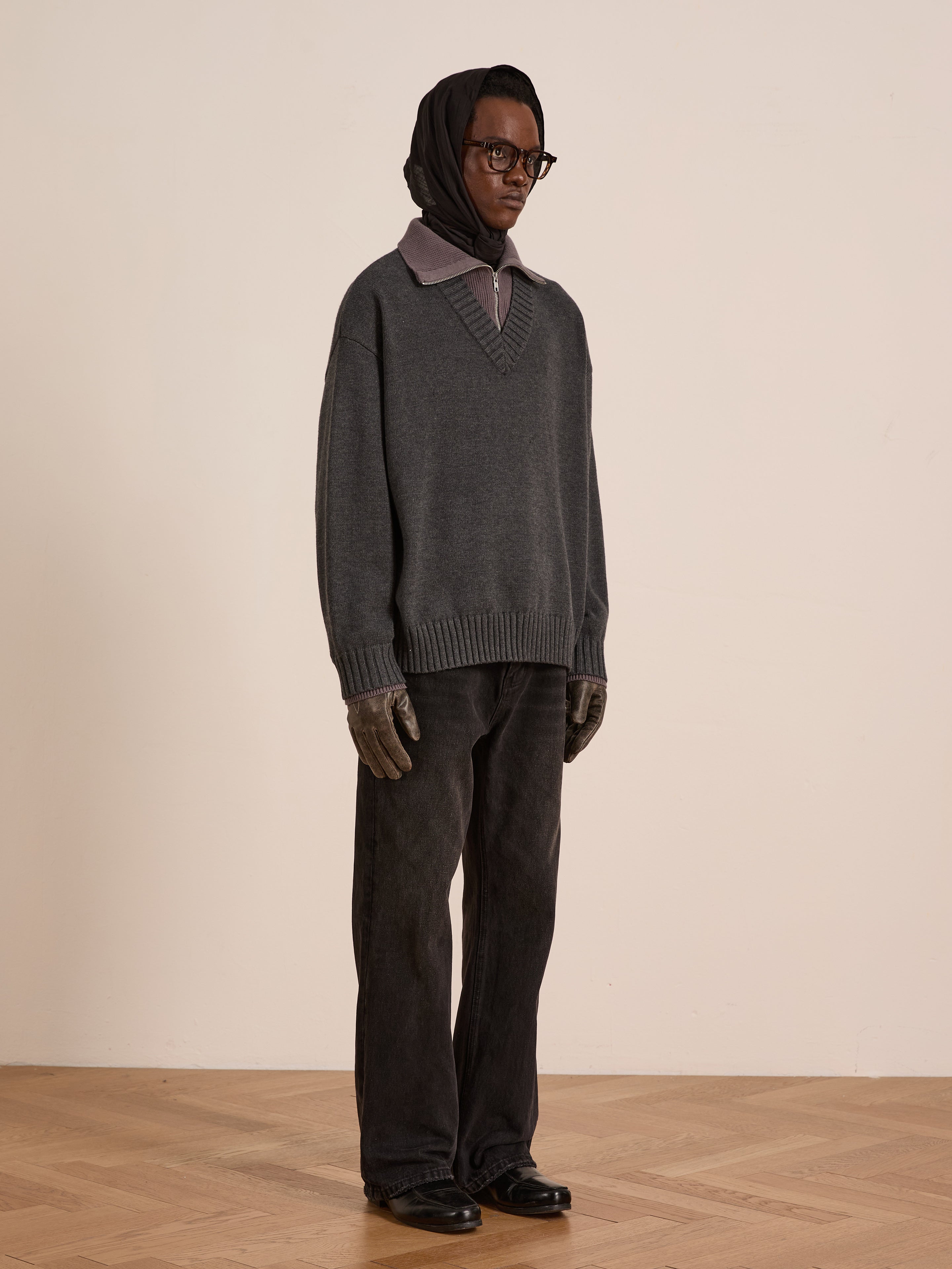 Indoors, a person wears the FOUND Double Layer Sweater over a collared shirt with a dark headscarf, glasses, gloves, loose dark pants, and black shoes—ideal cold-weather layering on a wooden floor against a plain wall.