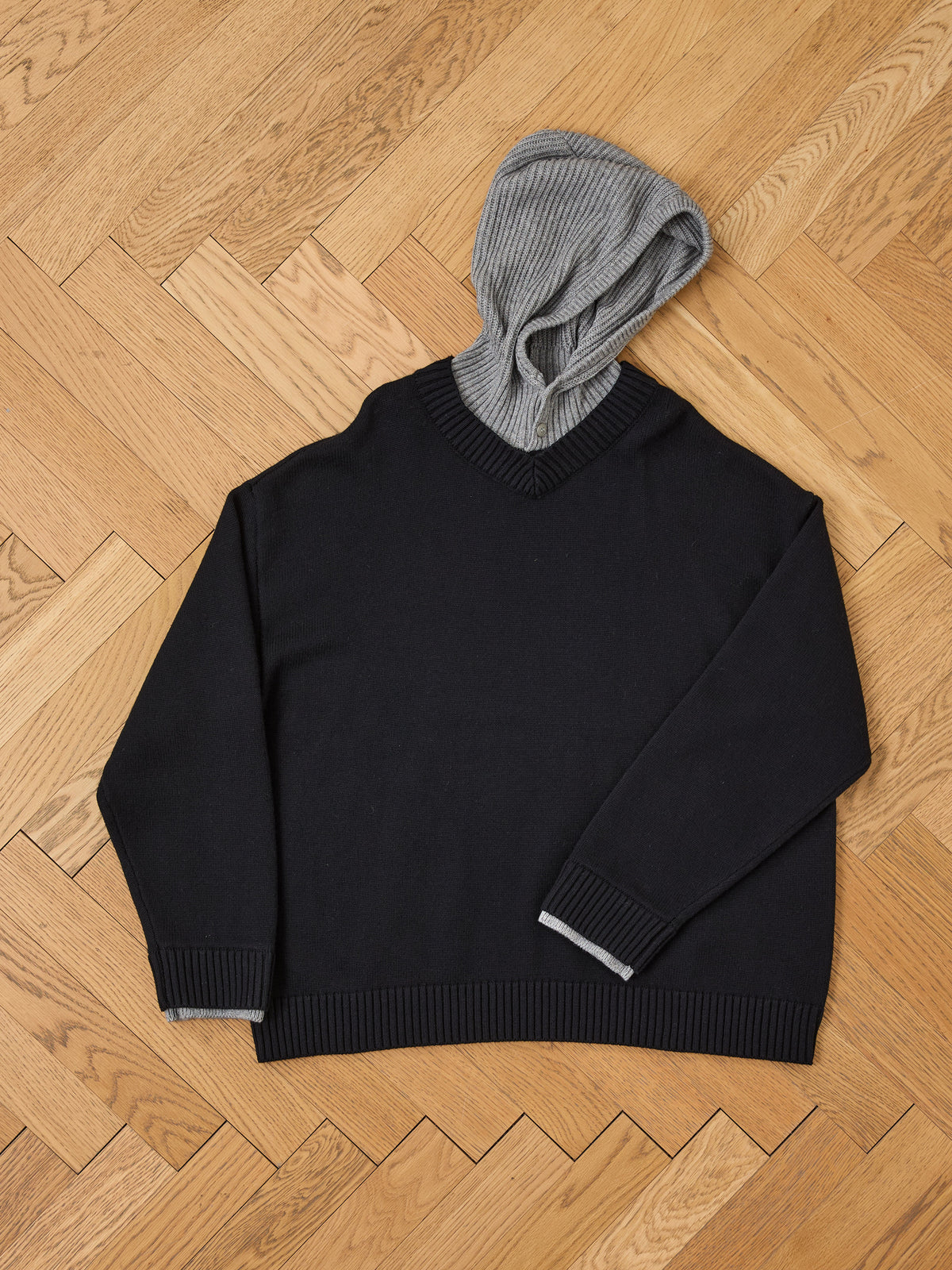 A black FOUND Double Layer V Neck Knit Sweater with a gray ribbed hood and gray cuffs is laid flat on a parquet wood floor.