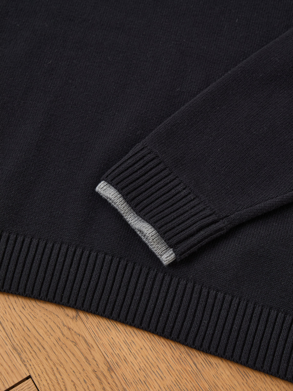 A close-up of the FOUND Double Layer V Neck Knit Sweater sleeve with a ribbed cuff and gray stripe, resting on a wooden surface.
