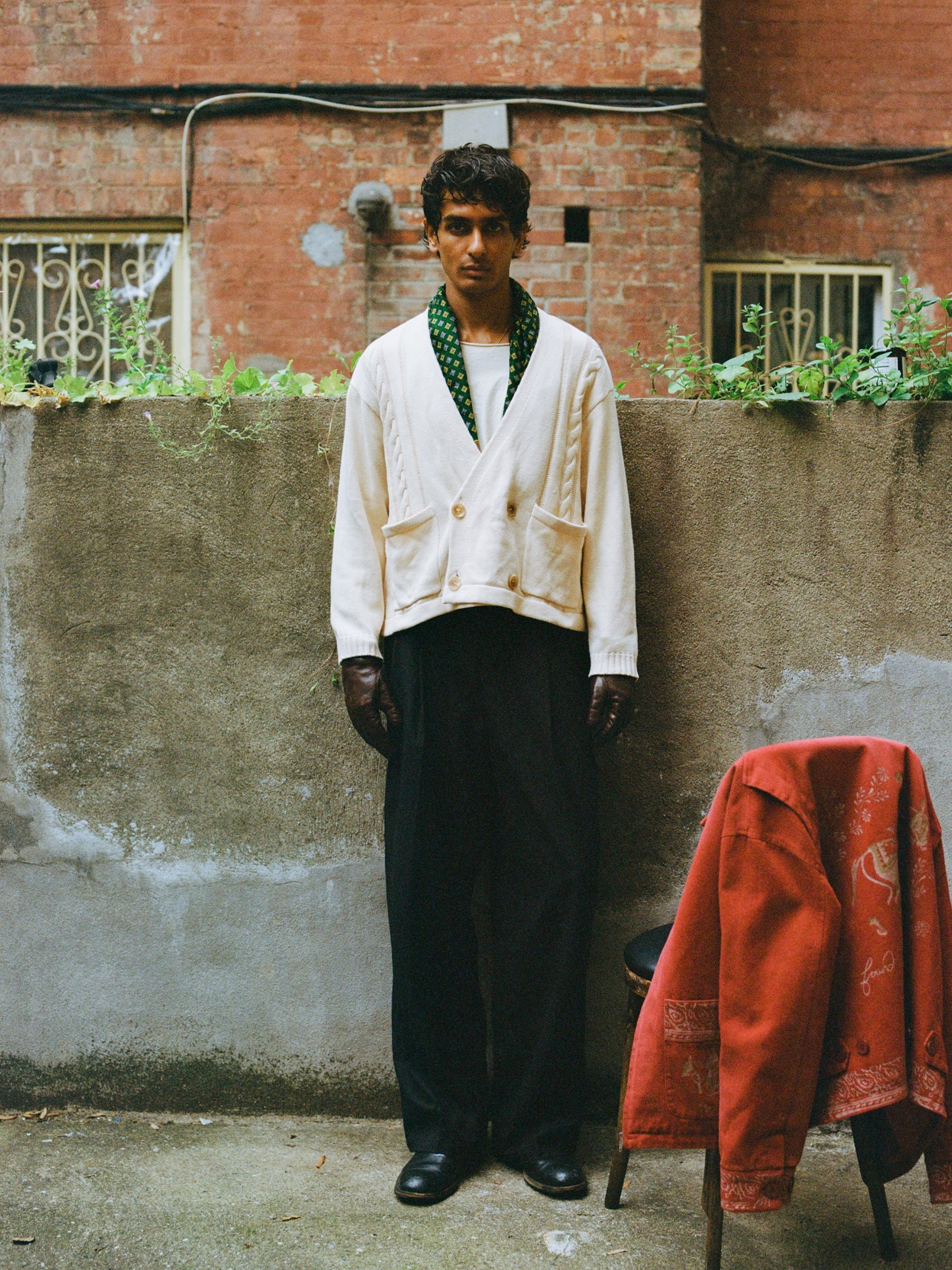 A man stands outdoors by a concrete wall, wearing the FOUND Double Breasted Knit Cardigan in white, paired with black trousers, a green scarf, and dark gloves; a red jacket rests on a nearby chair.