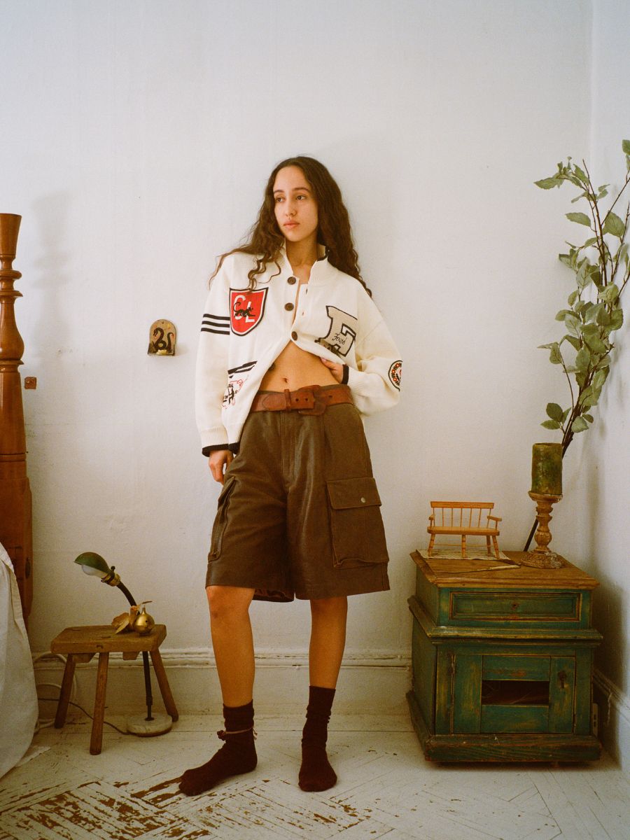 Amidst a utilitarian setting, an individual dons a white varsity jacket with FOUND's Dusk Distressed Genuine Leather Cargo Shorts. A small table, green cabinet, and plant enhance the scene's charm.