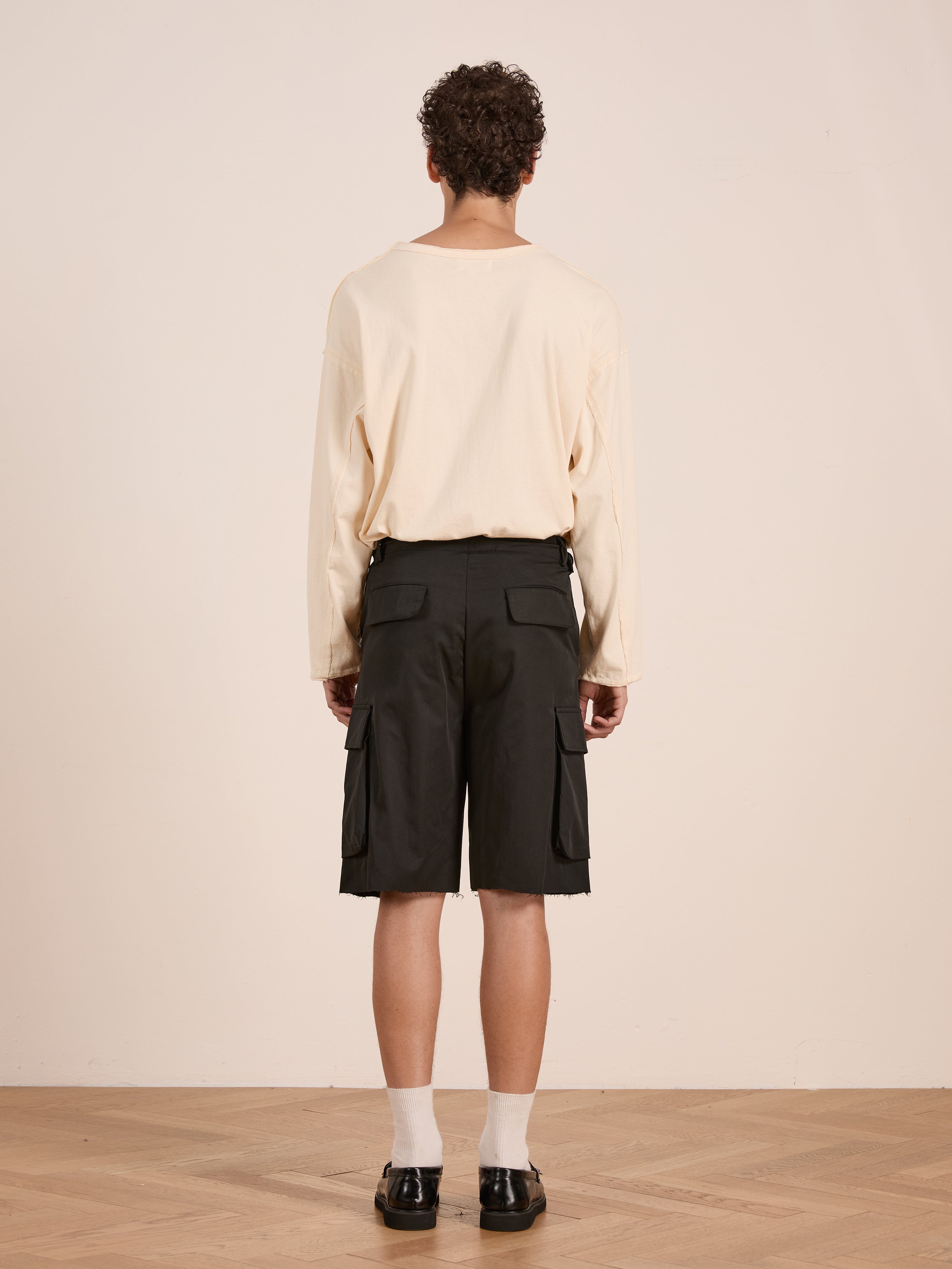 A person stands indoors on a wooden floor, facing away, wearing a long-sleeve beige shirt and Found Elbas Cargo Shorts with dual cargo pockets and adjustable waist tabs, paired with white socks and black shoes.