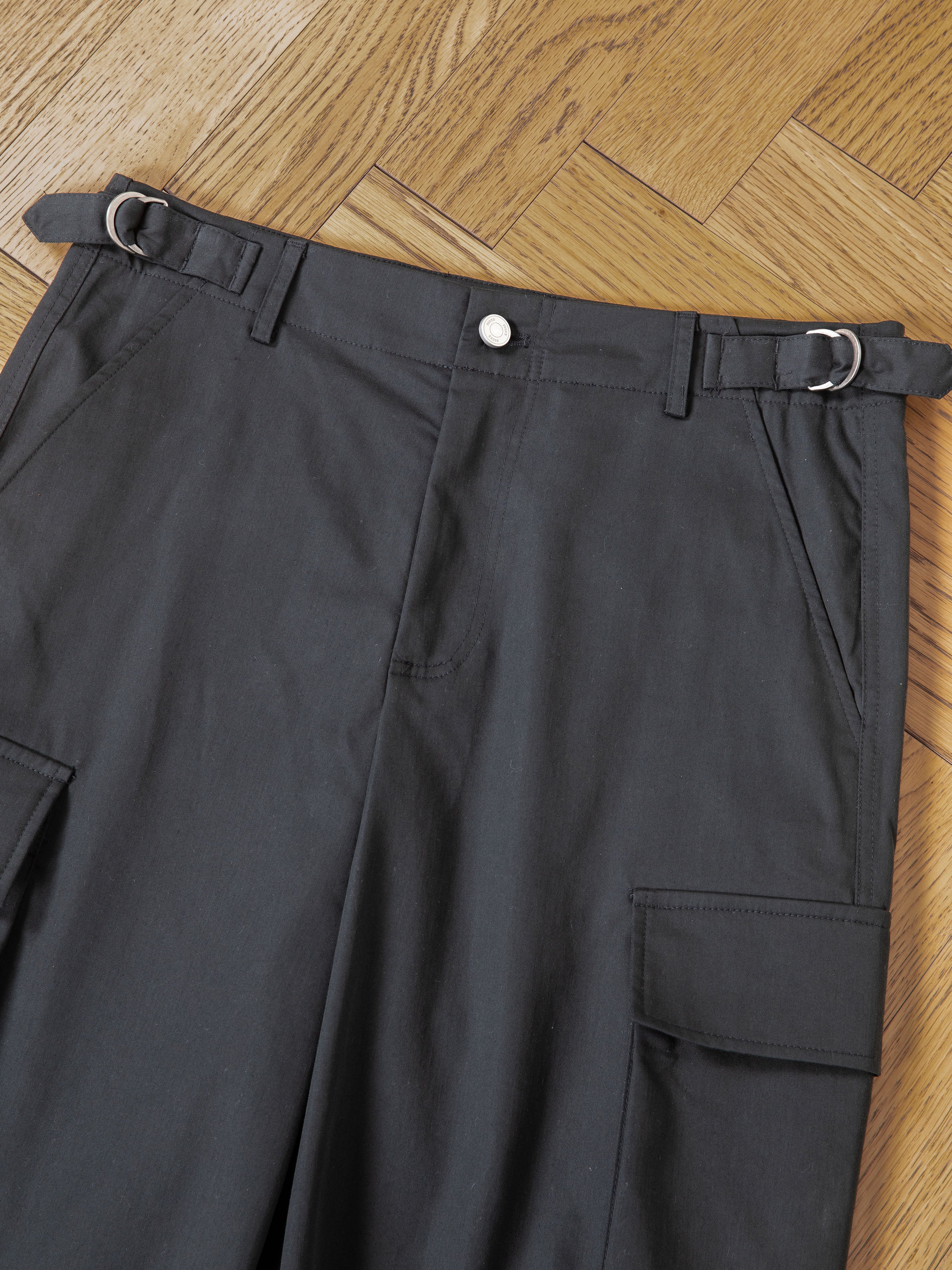 A pair of Found Elbas Cargo Shorts in dark gray, featuring dual cargo pockets, belt loops, and adjustable waist tabs with metal buckles, laid flat on a wooden parquet floor.