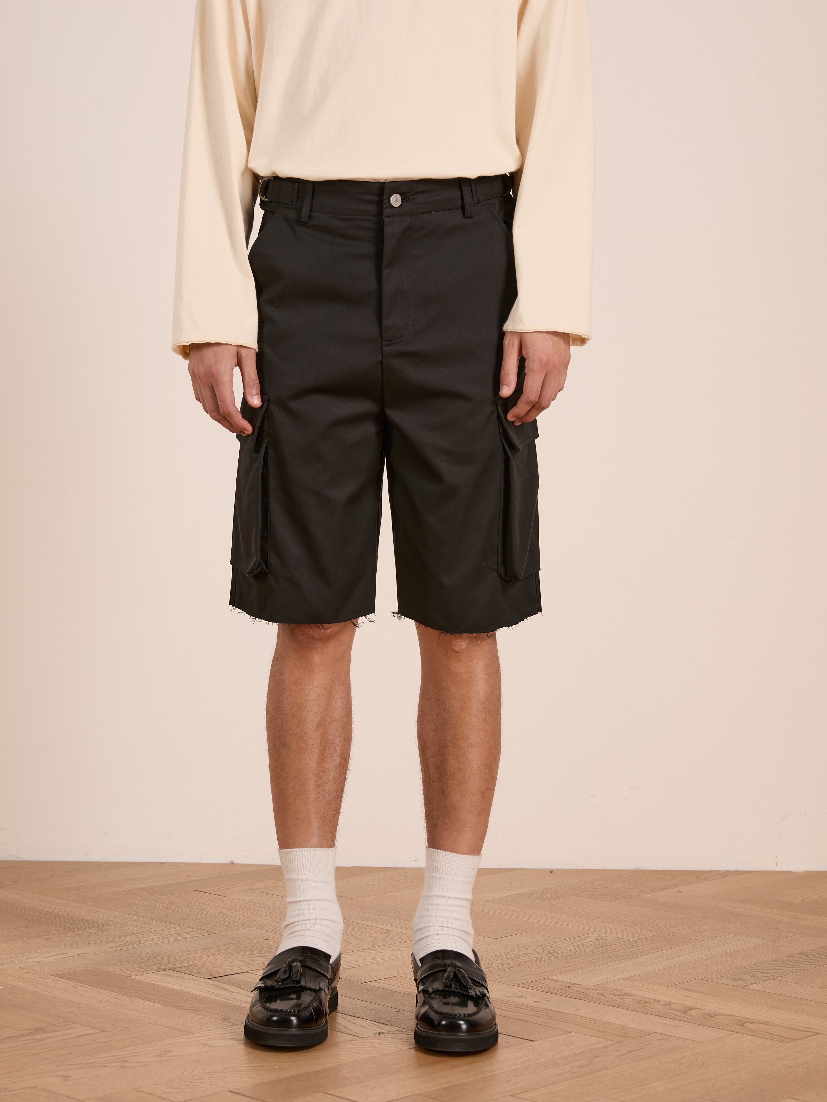 A person wearing a cream long-sleeve shirt, Found Elbas Cargo Shorts with dual cargo pockets and adjustable waist tabs, white socks, and black loafers stands on a wooden floor against a plain background.