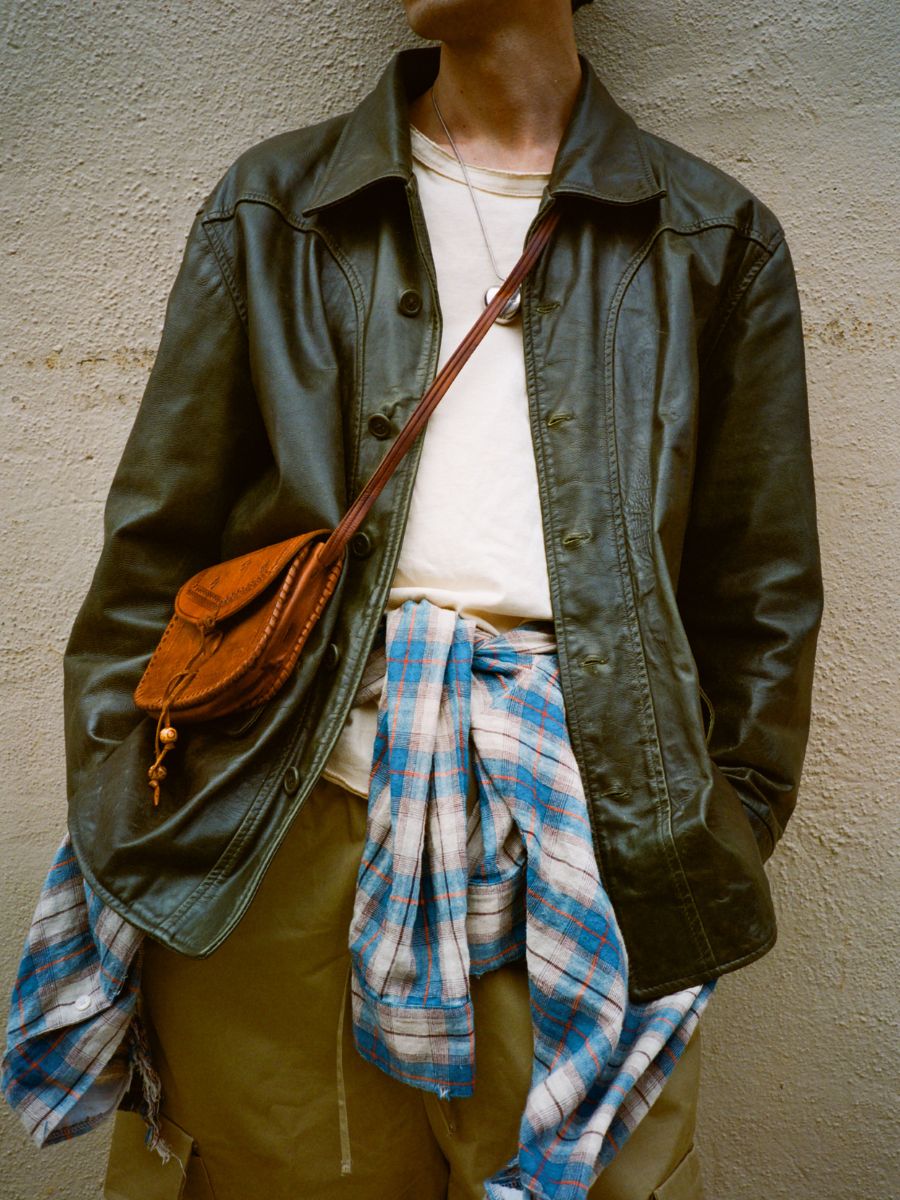 Individual wearing the FOUND Leather Field Jacket, a white shirt, plaid shirt tied at the waist, khaki pants, and a brown crossbody bag, standing against a textured beige wall.