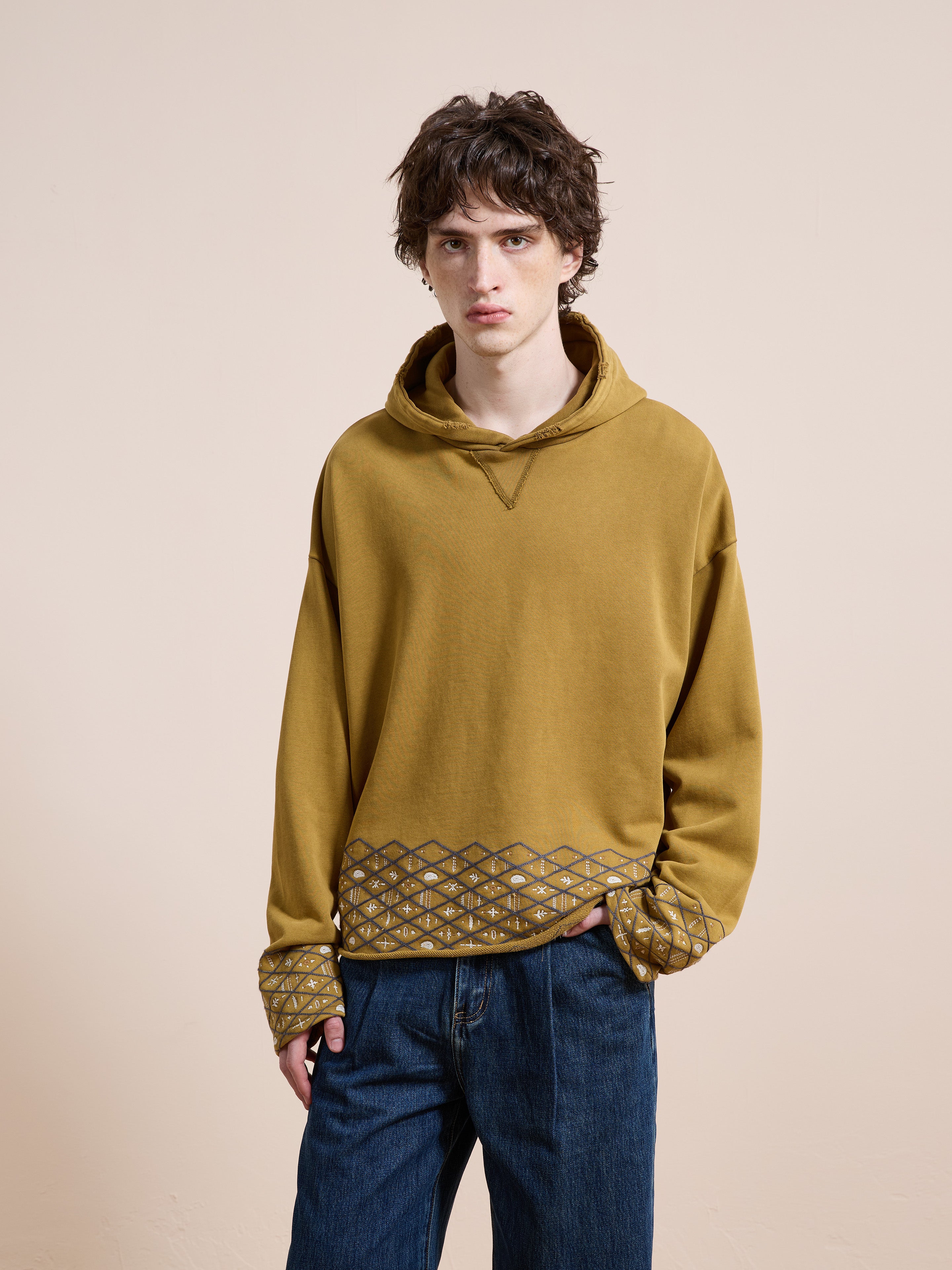 A person with short brown hair wears the FOUND Embroidered Border Hoodie in mustard yellow with geometric patterns, paired with blue jeans, standing against a plain beige background.