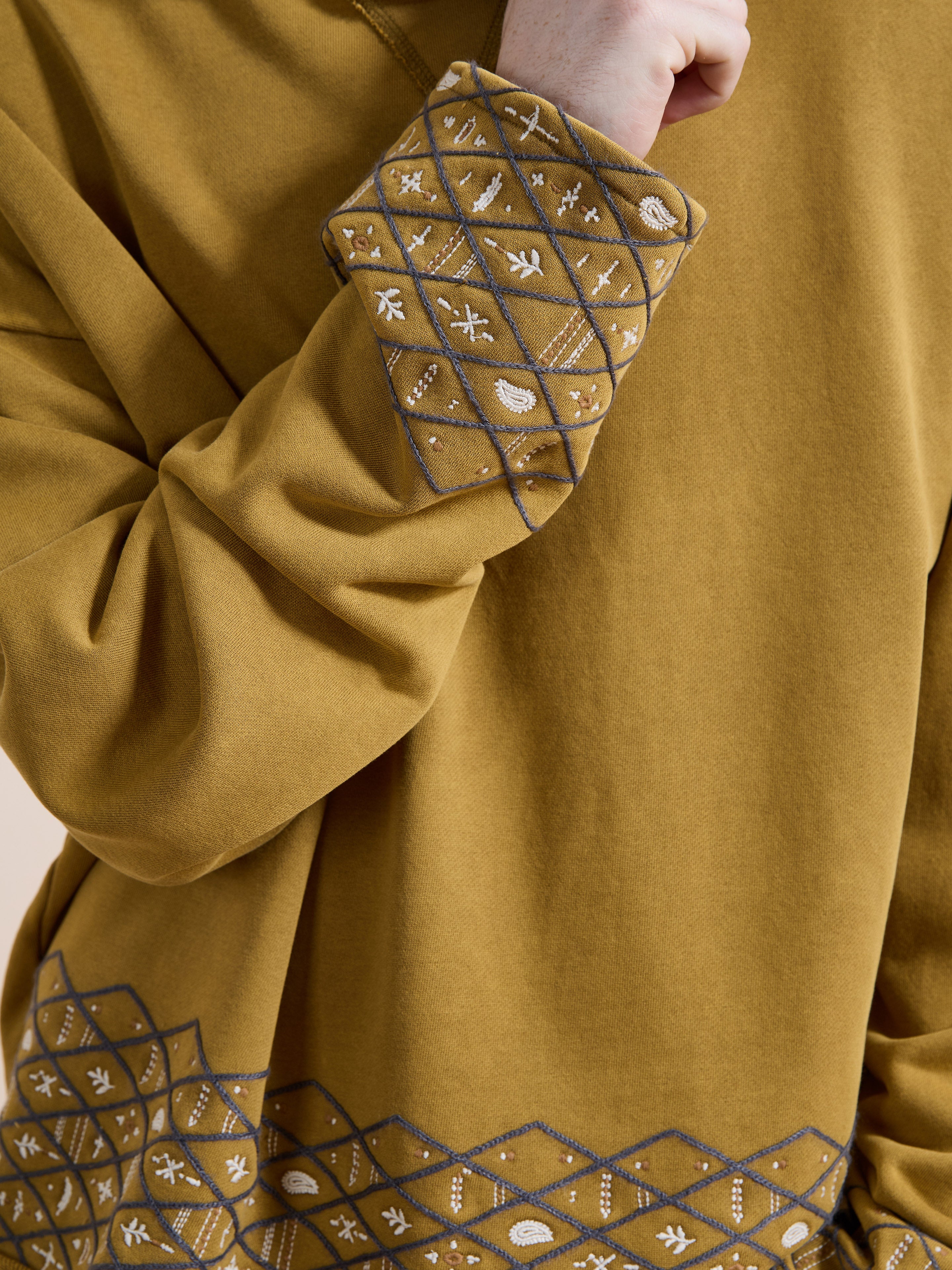 A person wearing the FOUND Embroidered Border Hoodie in mustard, featuring geometric and floral embroidery on the cuffs and hem, holds their hand near their face.