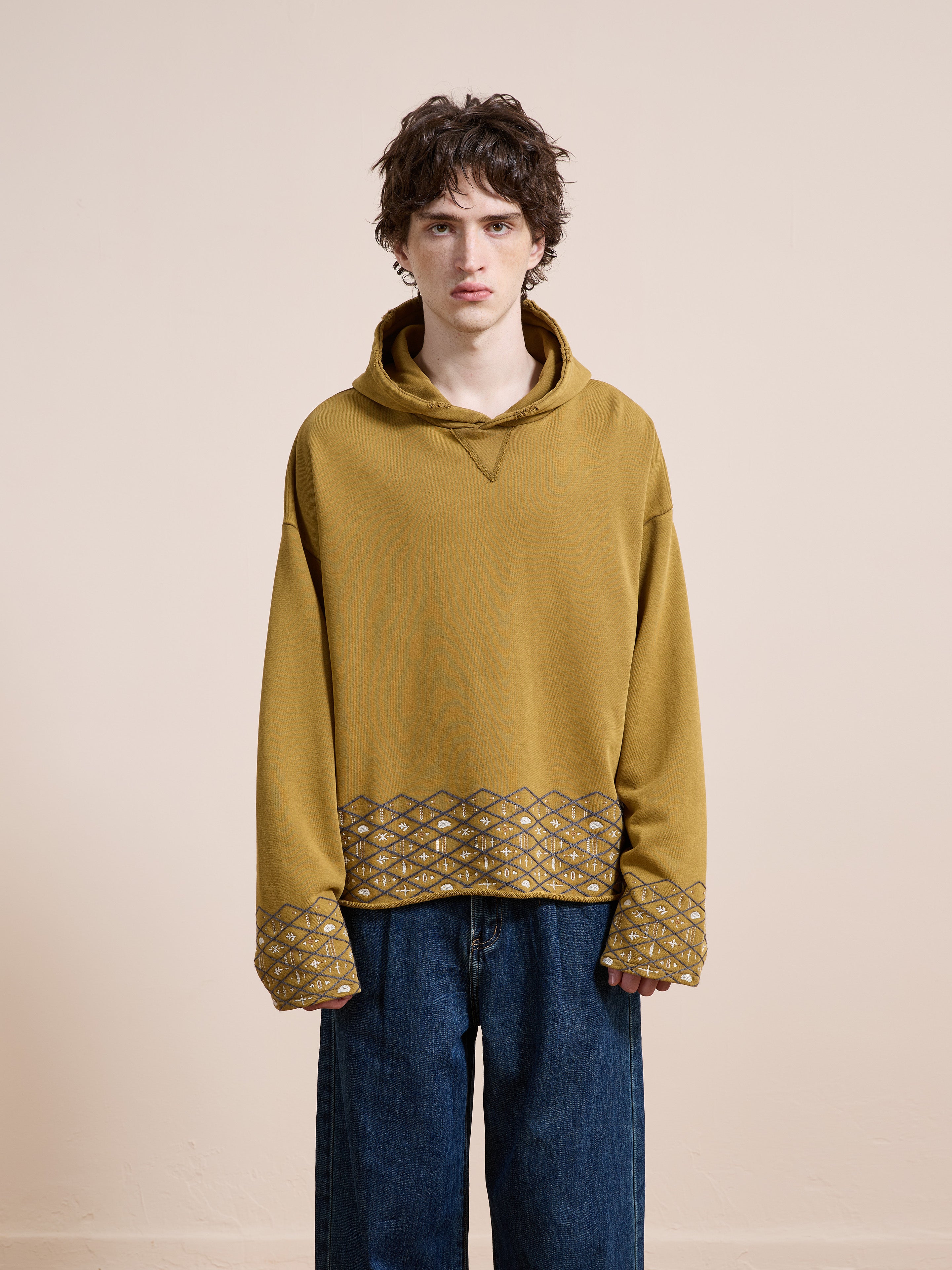 A person stands against a plain light background wearing the FOUND Embroidered Border Hoodie in mustard yellow, featuring patterned cuffs and hem, paired with wide-leg blue jeans.