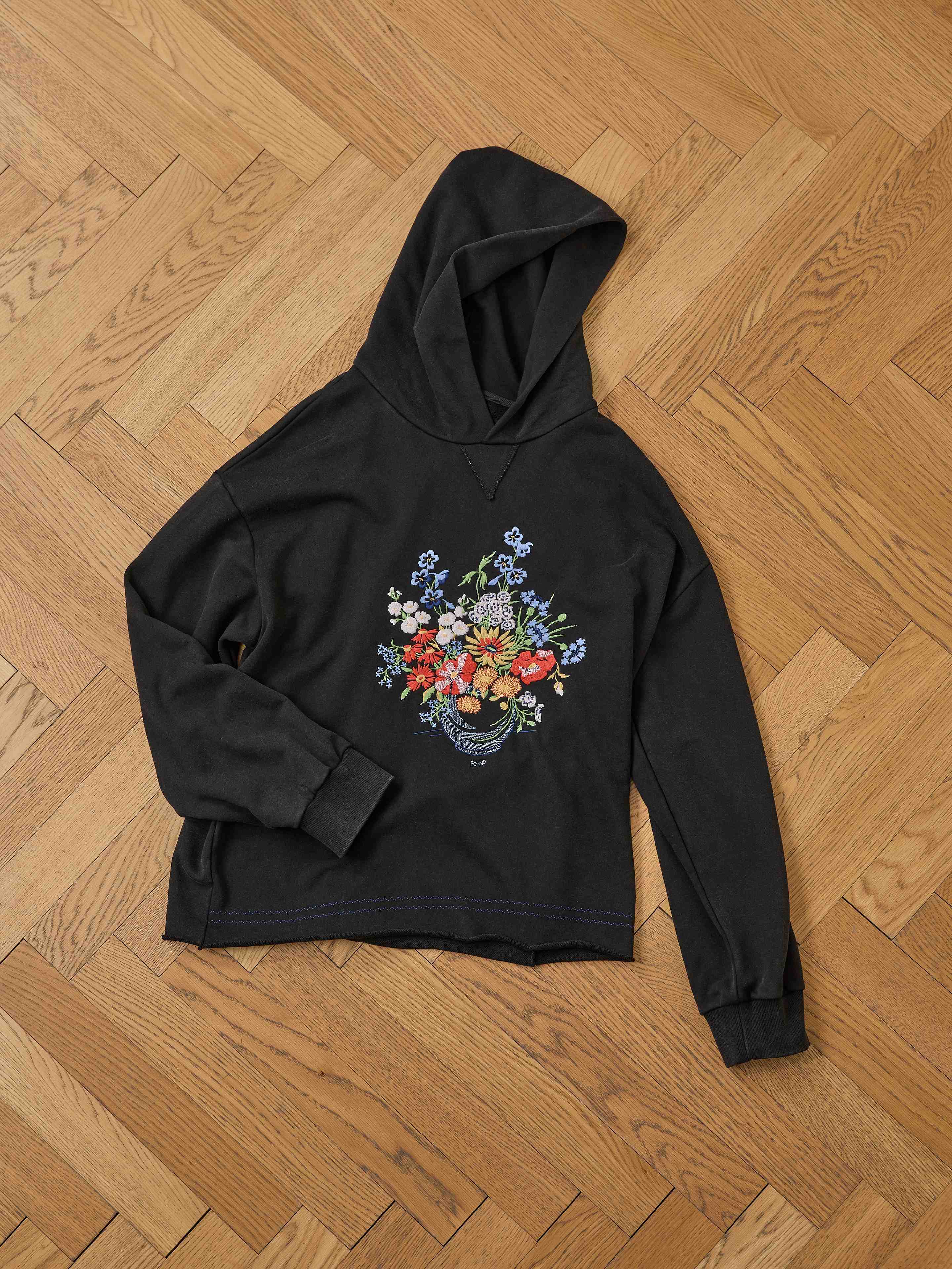 The FOUND Bouquet Flowers Hoodie in black features colorful floral embroidery, displayed flat on a wooden herringbone floor to showcase its vibrant bouquet design.