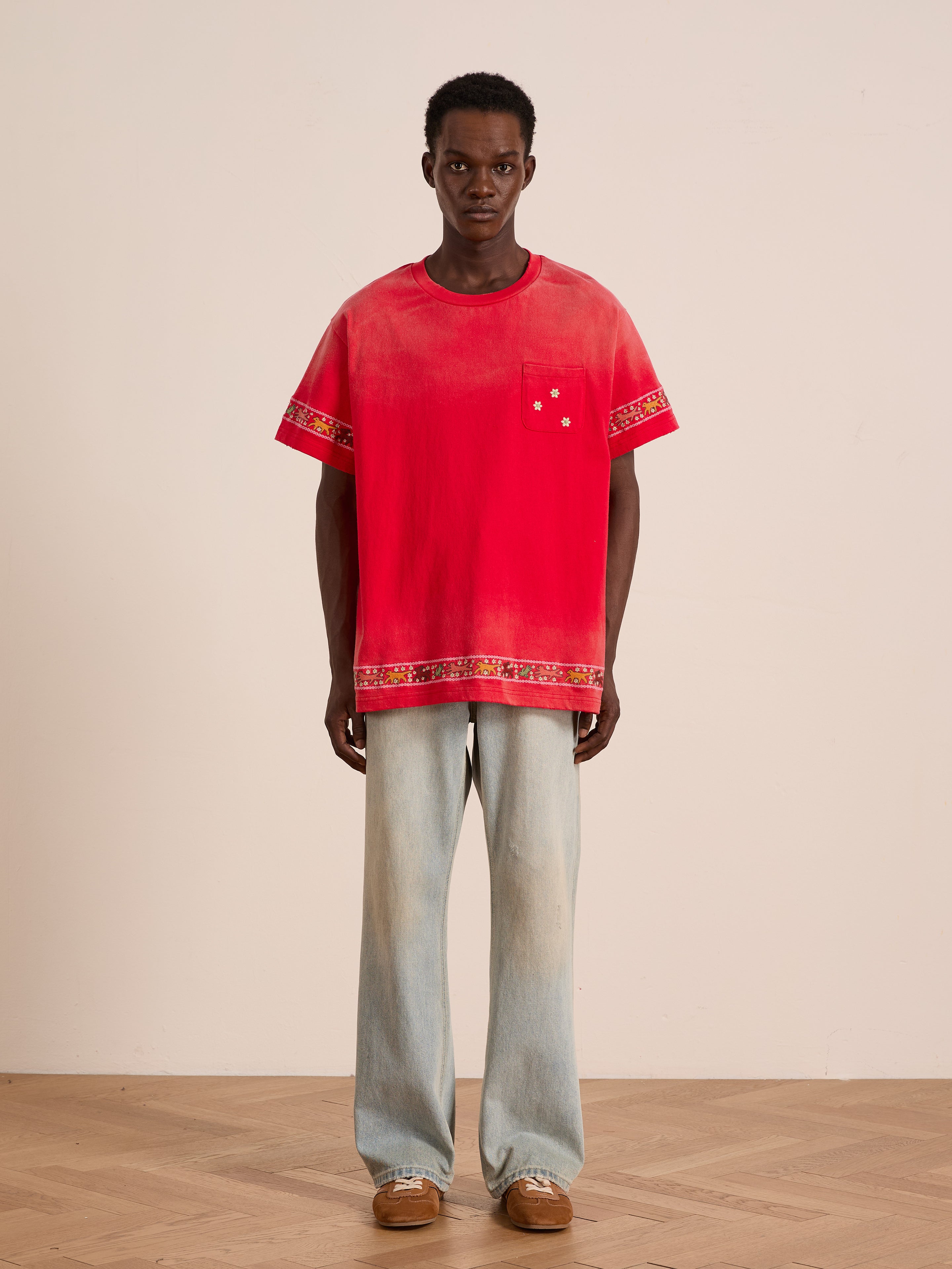 A person stands indoors against a plain background wearing the FOUND Horse Motif Embroidered Tee, featuring red embroidery and decorative trim, paired with light blue wide-leg jeans and brown shoes.
