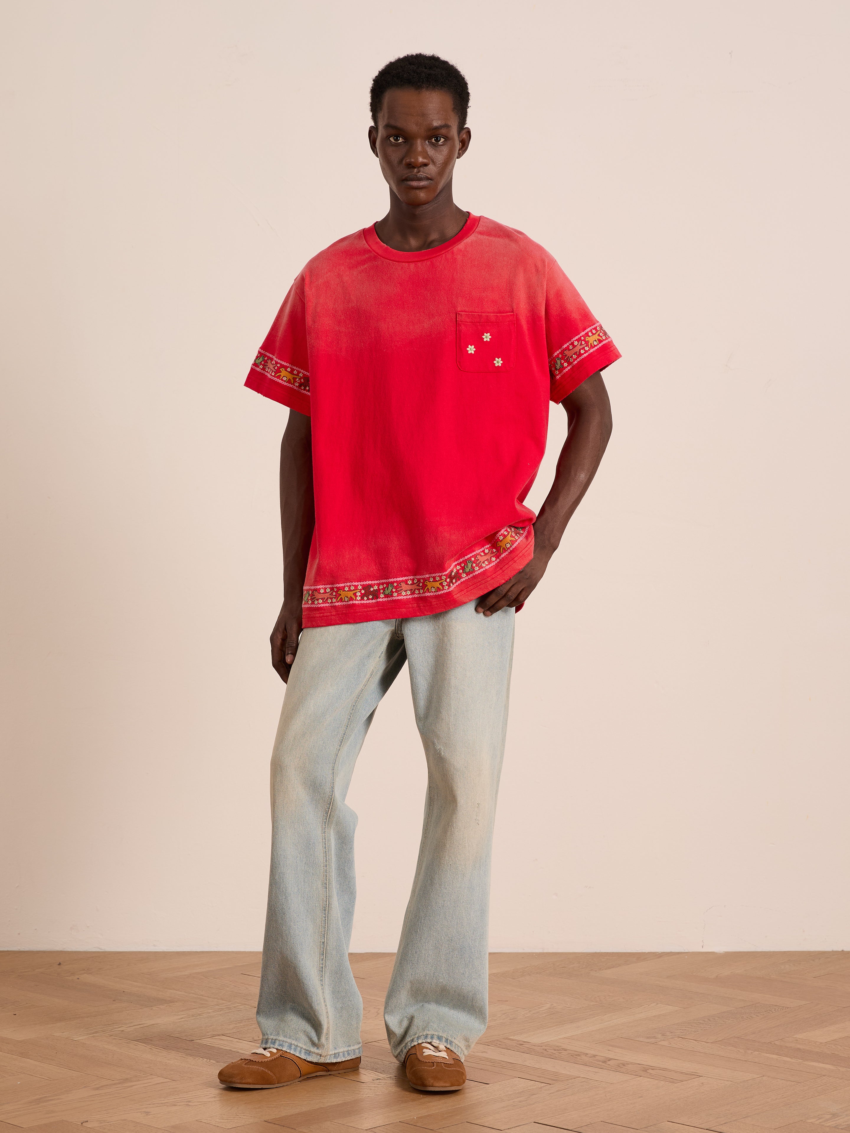 A man stands indoors wearing FOUND’s Horse Motif Embroidered Tee in red, paired with light blue jeans and brown shoes on a wooden floor against a plain wall.