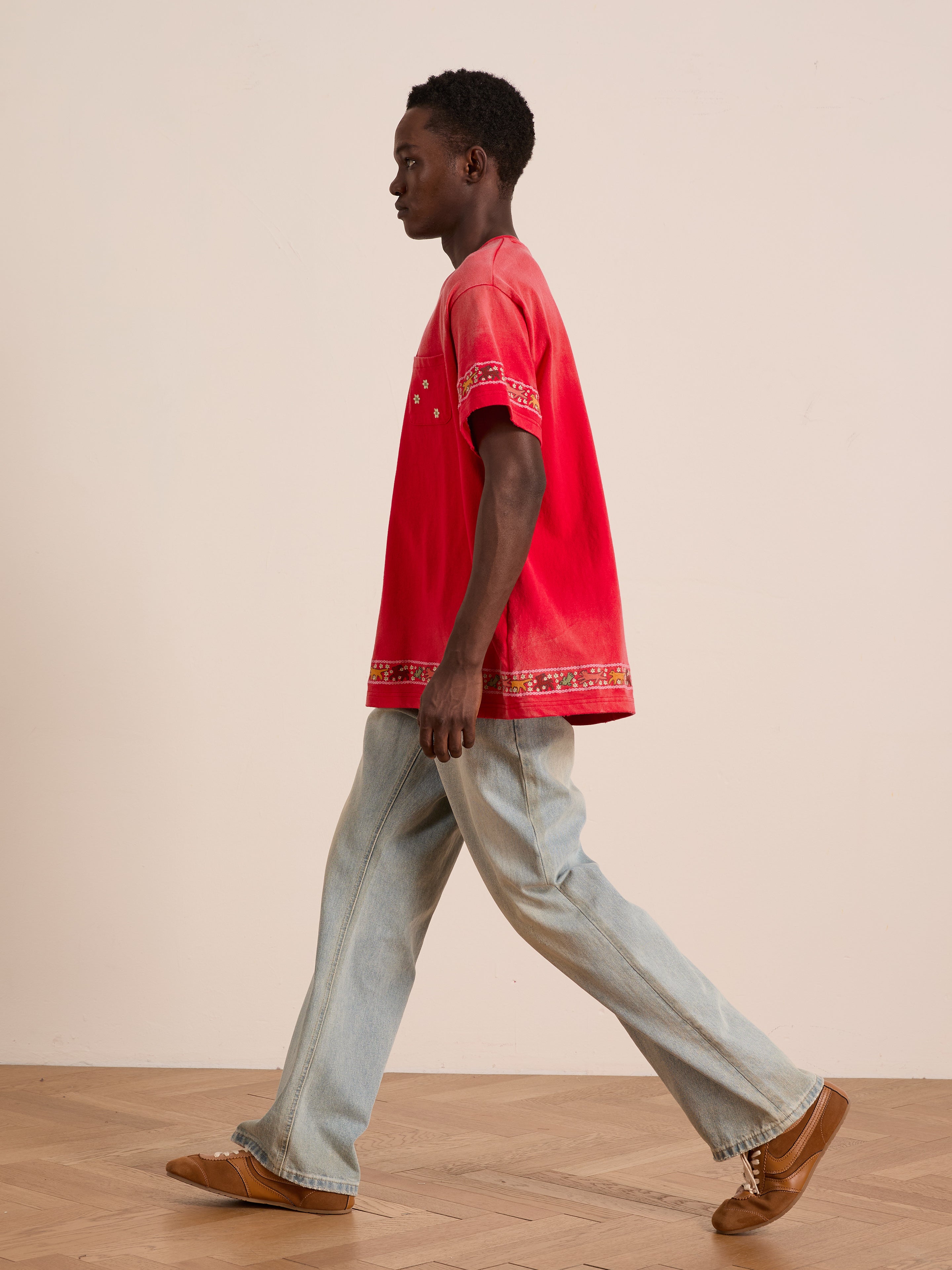 A man walks indoors against a plain background wearing the FOUND Horse Motif Embroidered Tee, a unisex relaxed-fit red shirt, paired with light blue jeans and brown shoes.
