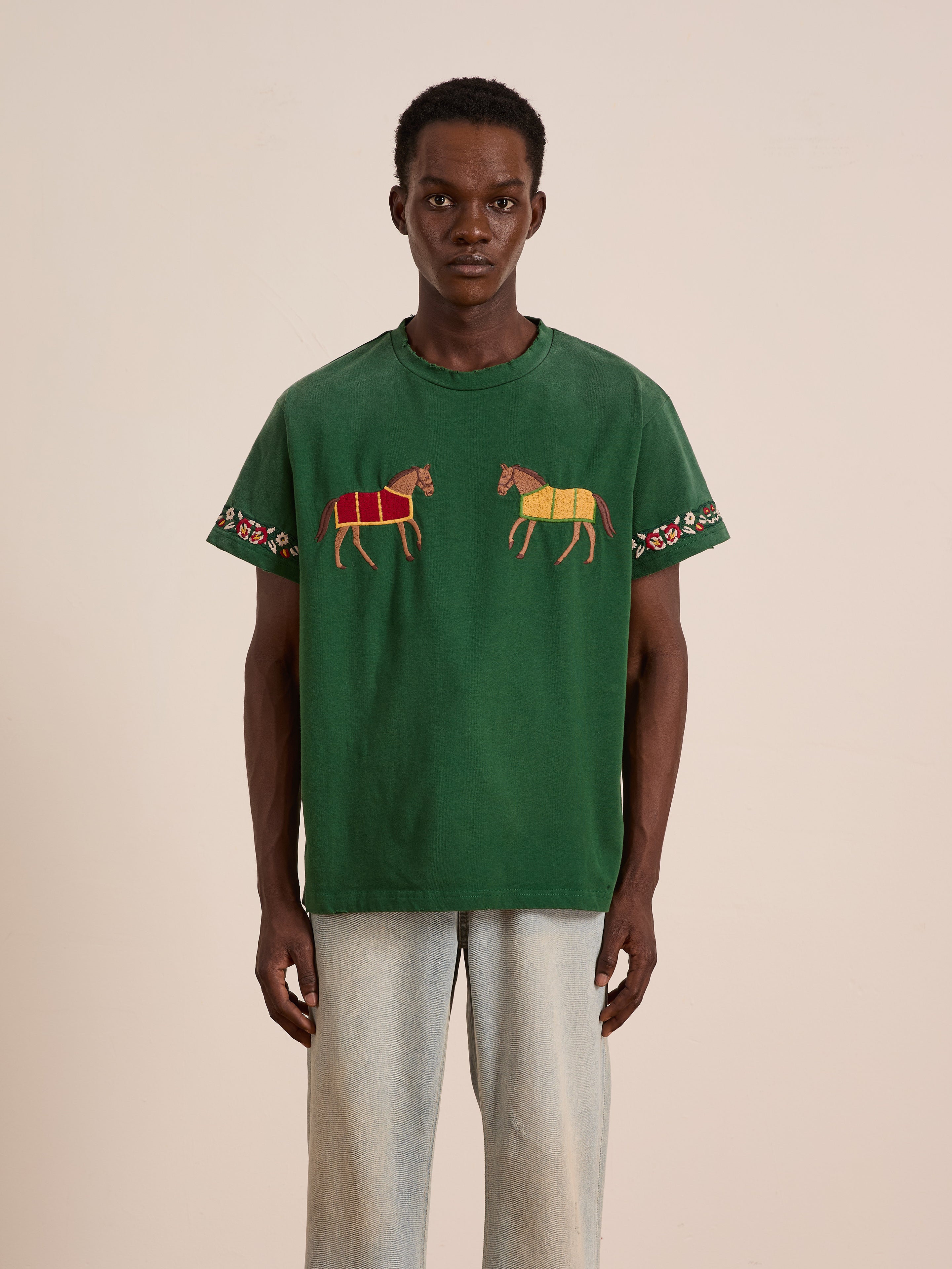 Someone models the FOUND Horse Equine Tee, featuring green short sleeves with embroidered horses on the front and floral embroidery on the sleeves, paired with light blue jeans against a plain background.