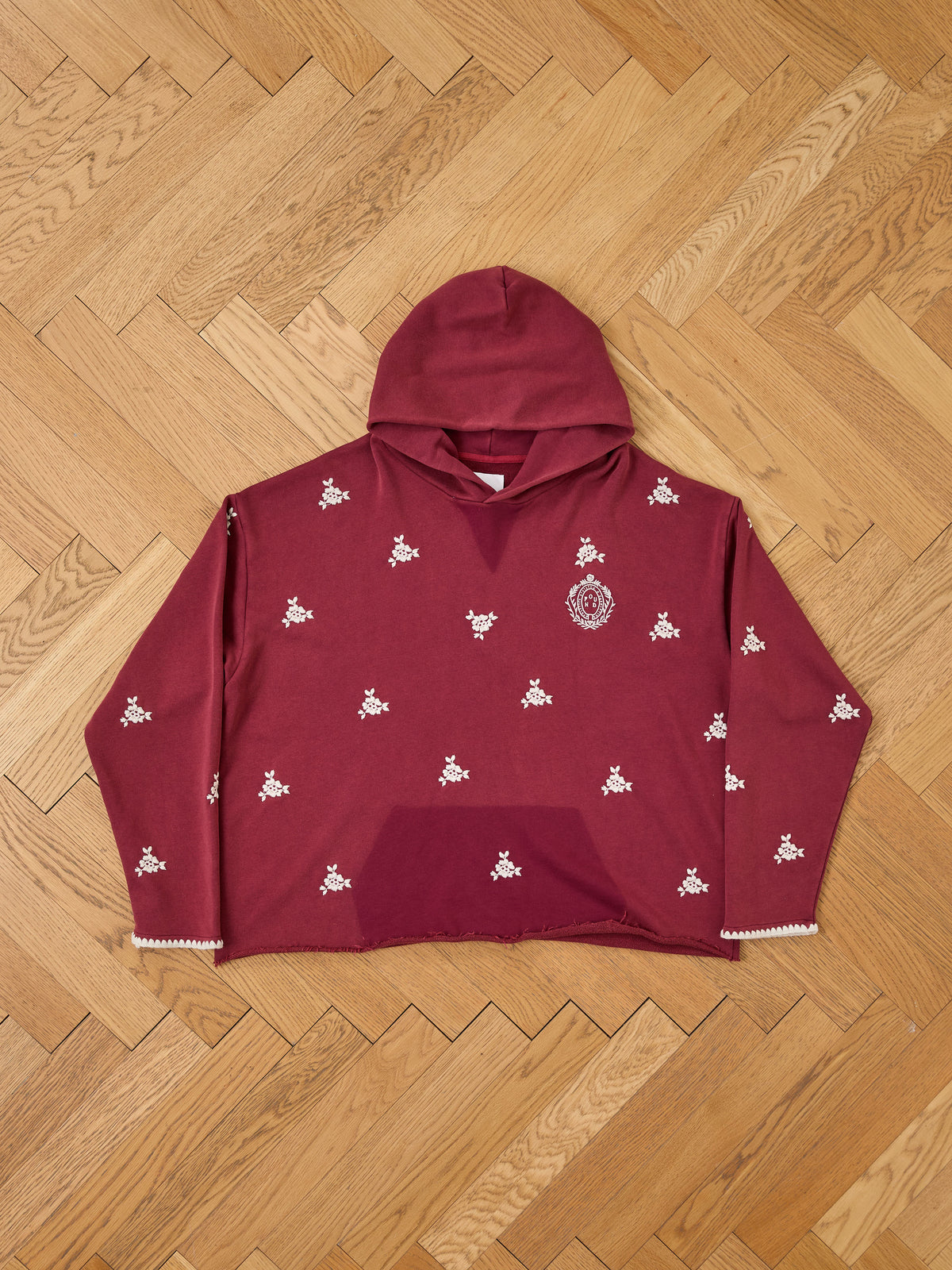 FOUND Embroidered Motif Hoodie in maroon, unisex relaxed fit with white floral embroidery on the chest and a front pocket, shown laid flat on a herringbone floor.
