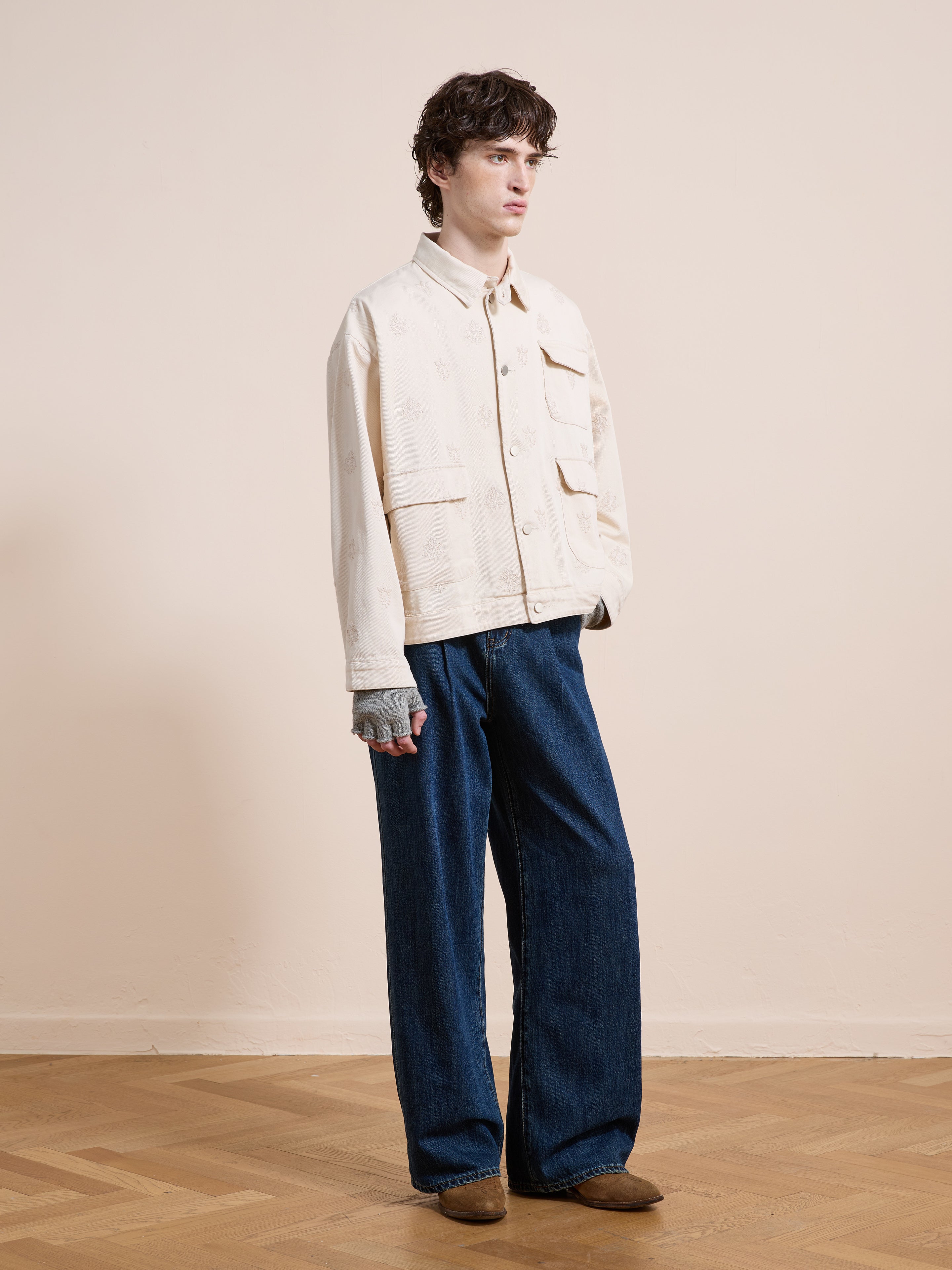 A person stands indoors on a wooden floor against a beige wall wearing the Found Embroidered Motif Jacket, dark wide-leg jeans, brown shoes, and fingerless gloves.