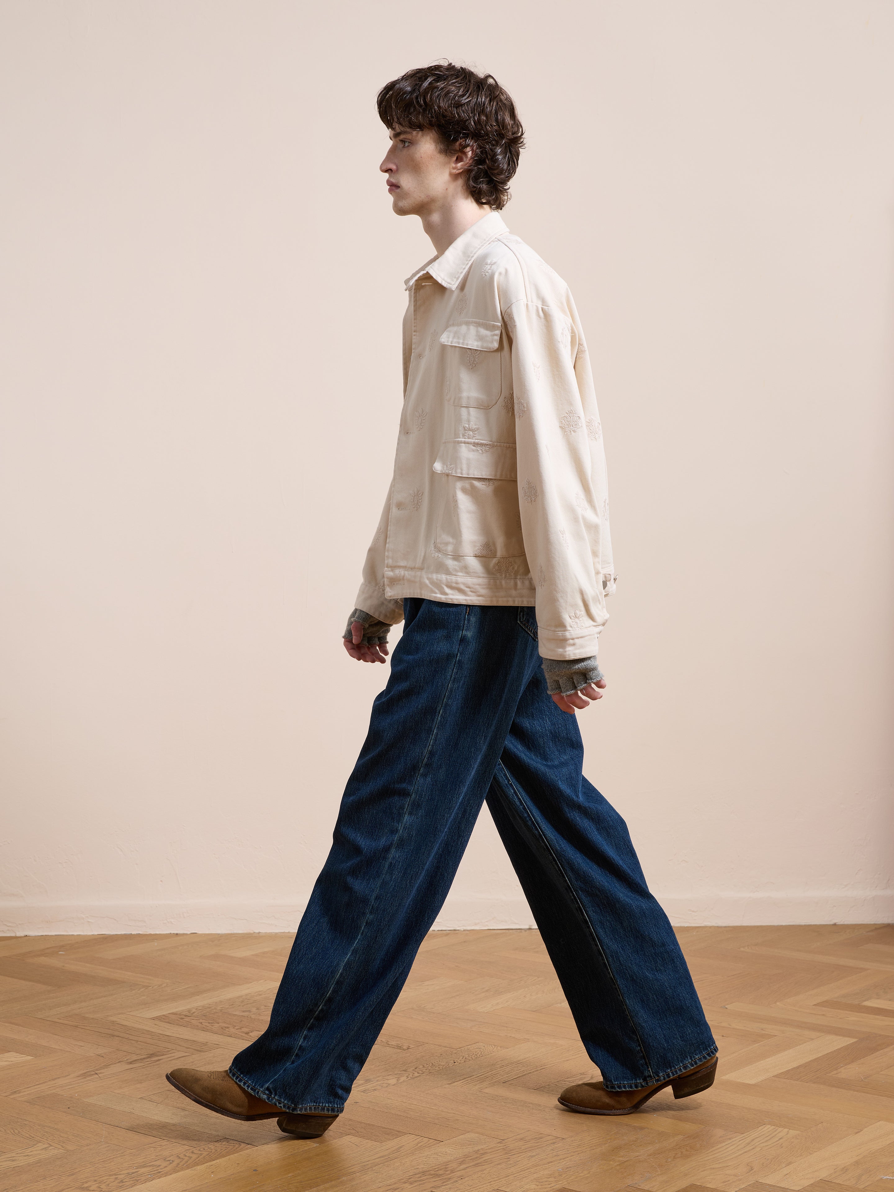 A person with curly hair, wearing the Found Embroidered Motif Jacket and wide-leg blue jeans with brown shoes, walks indoors on a wooden floor against a beige wall.