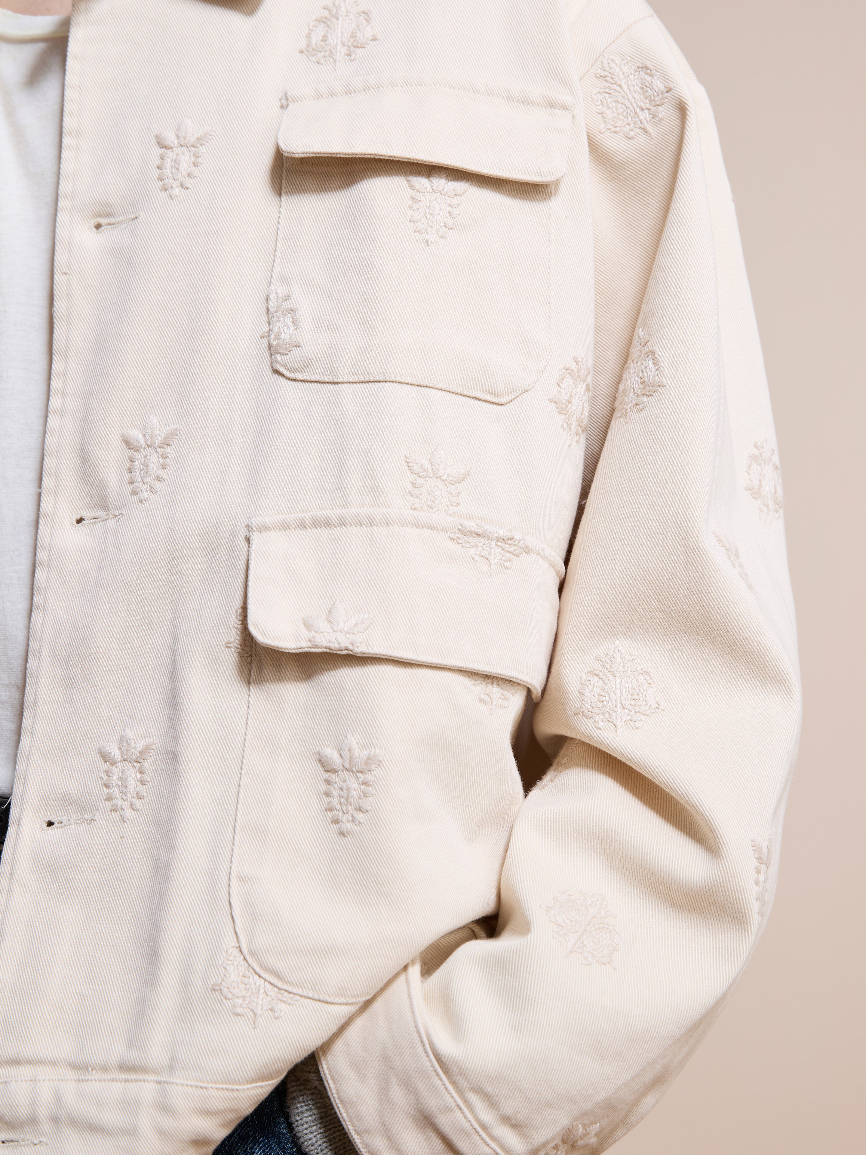 A close-up of Found's cream Embroidered Motif Jacket with front pockets and floral motifs, layered over a white shirt. This unisex jacket offers a relaxed fit for effortless style.