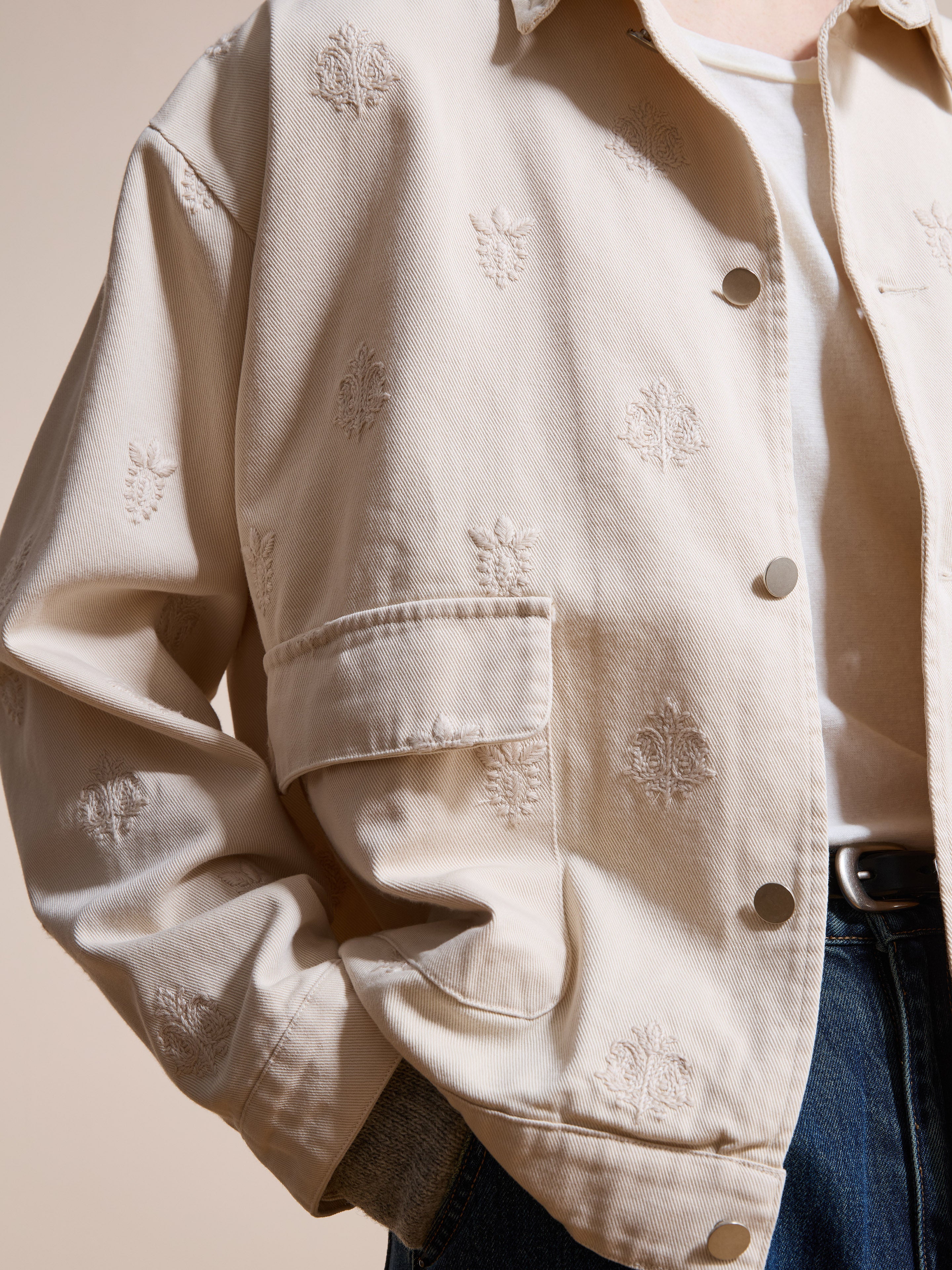 A person wears the Found Embroidered Motif Jacket in light beige over a white shirt and blue jeans, with one hand in their pocket.