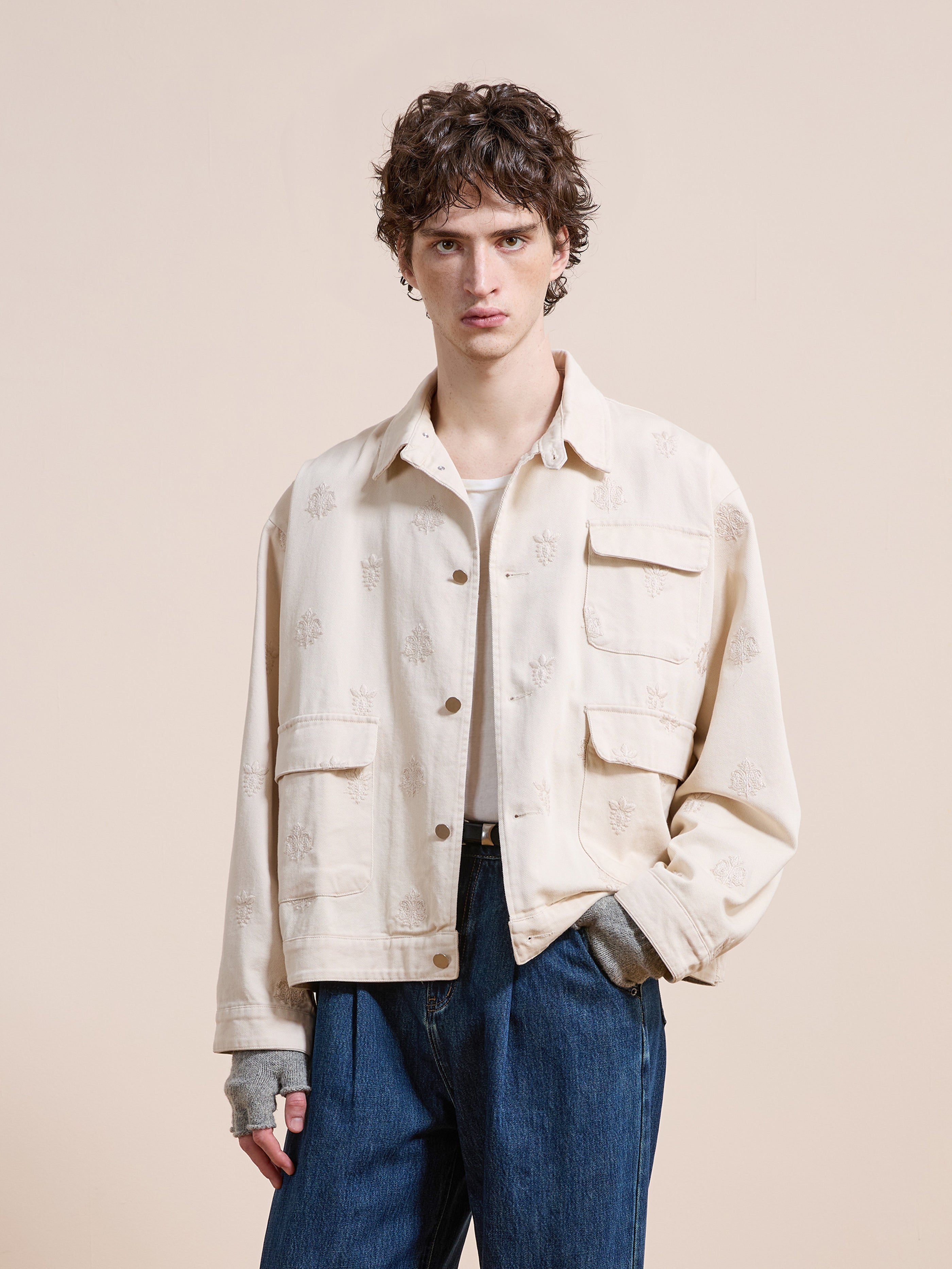 A young person with curly hair wears the Found Embroidered Motif Jacket in a unisex relaxed fit, featuring two front pockets, over blue jeans while standing against a plain beige background.