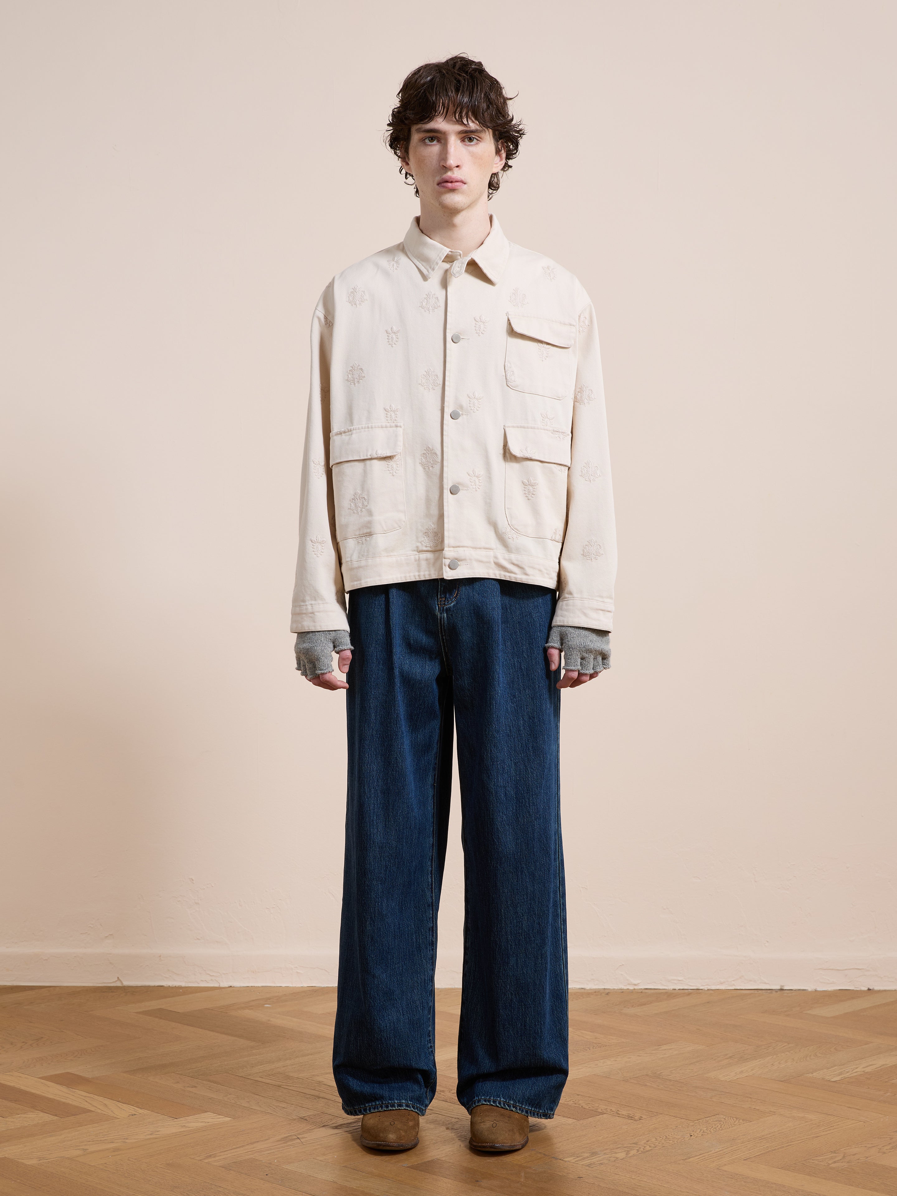 Person stands indoors against a plain wall, wearing Found's Embroidered Motif Jacket with relaxed fit, layered gray sleeves, wide-leg blue jeans, and brown shoes on a wooden floor.