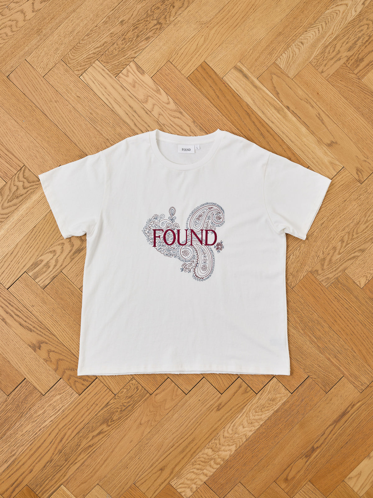 The Embroidered Paisley Tee by FOUND is a white unisex cotton shirt with paisley print and bold red “FOUND” on the front, shown on a wooden herringbone floor.