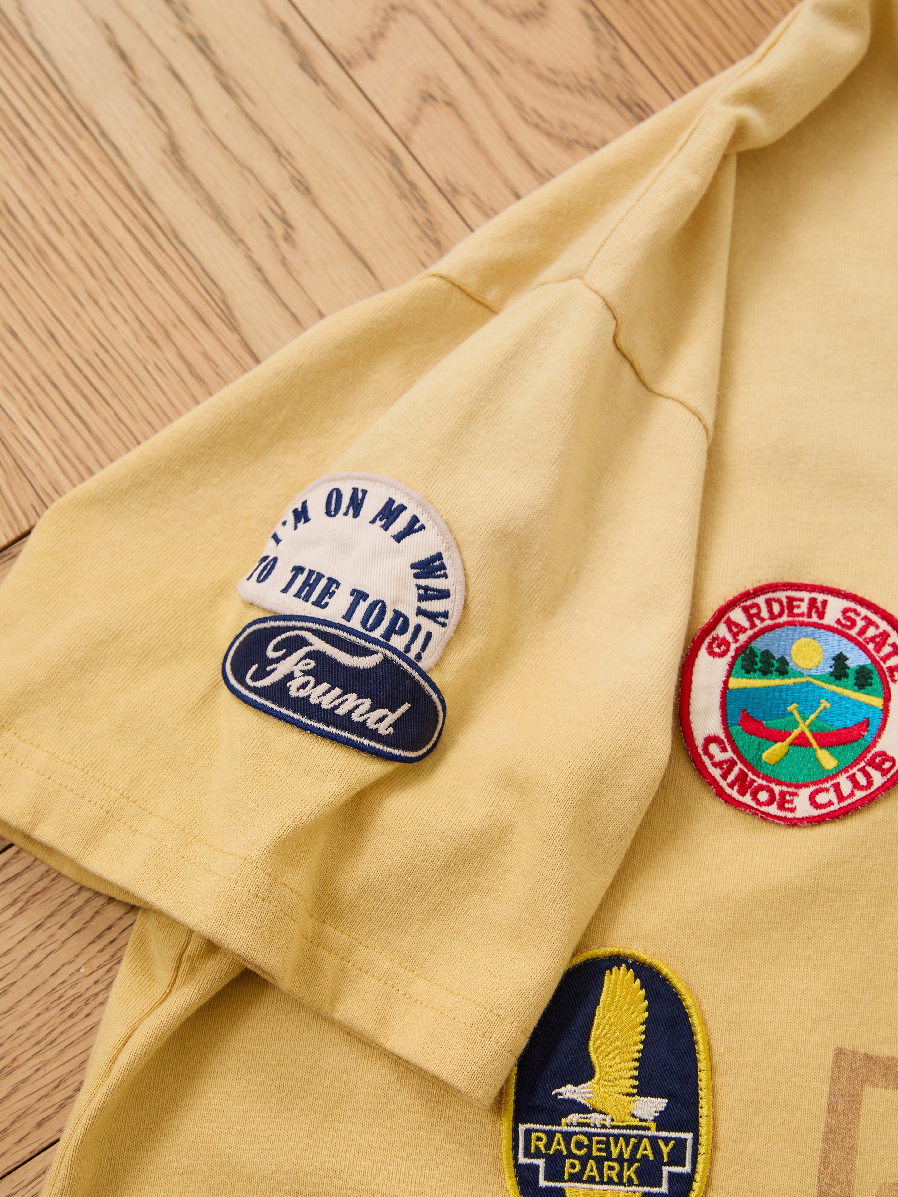 The FOUND Patchwork Camp Tee is a unisex, relaxed fit yellow tee with vintage-inspired embroidered patches such as "I'M ON MY WAY TO THE TOP!!" and "RACEWAY PARK," shown on a wooden surface.