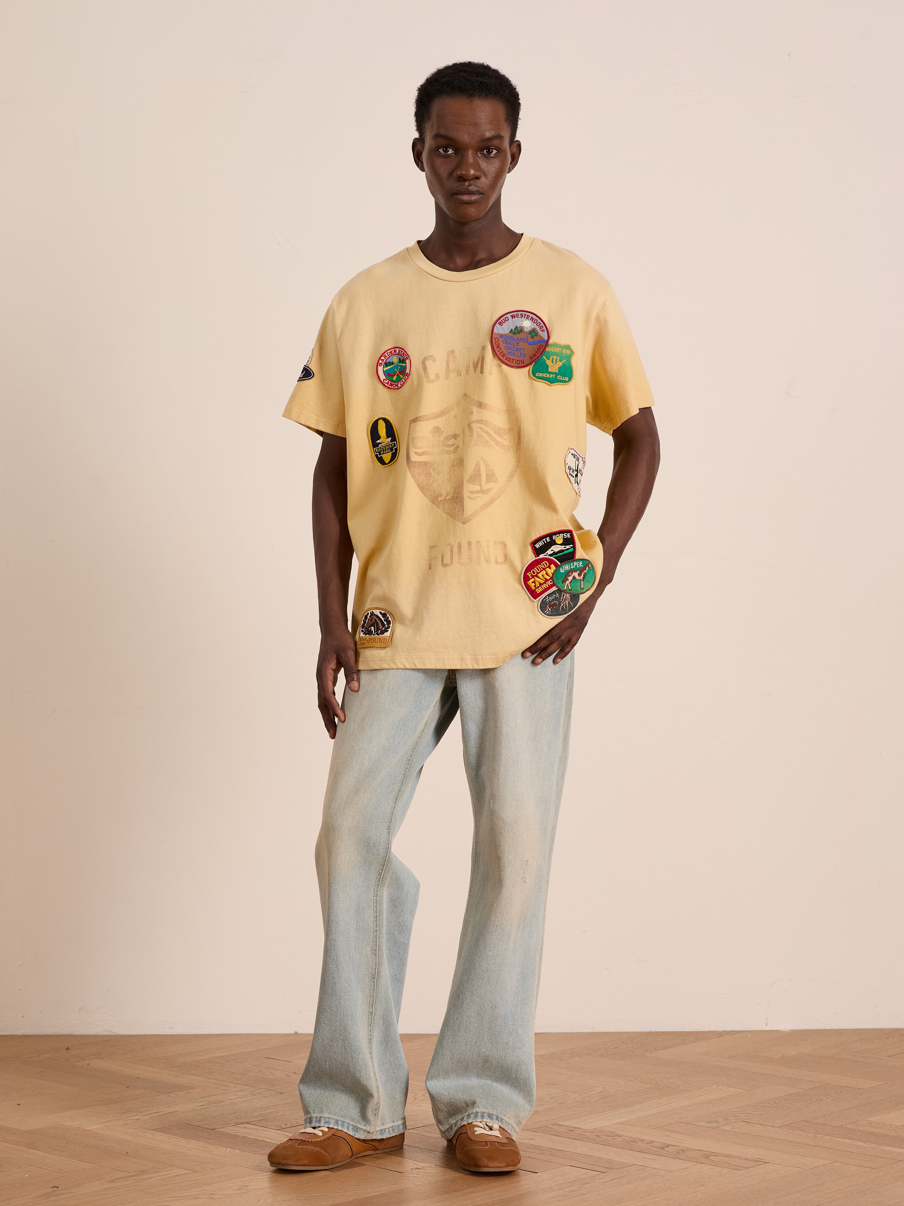 Indoors, a person wears the FOUND Patchwork Camp Tee in pale yellow with vintage-inspired embroidered patches, paired with light blue jeans and brown shoes, standing against a plain background.