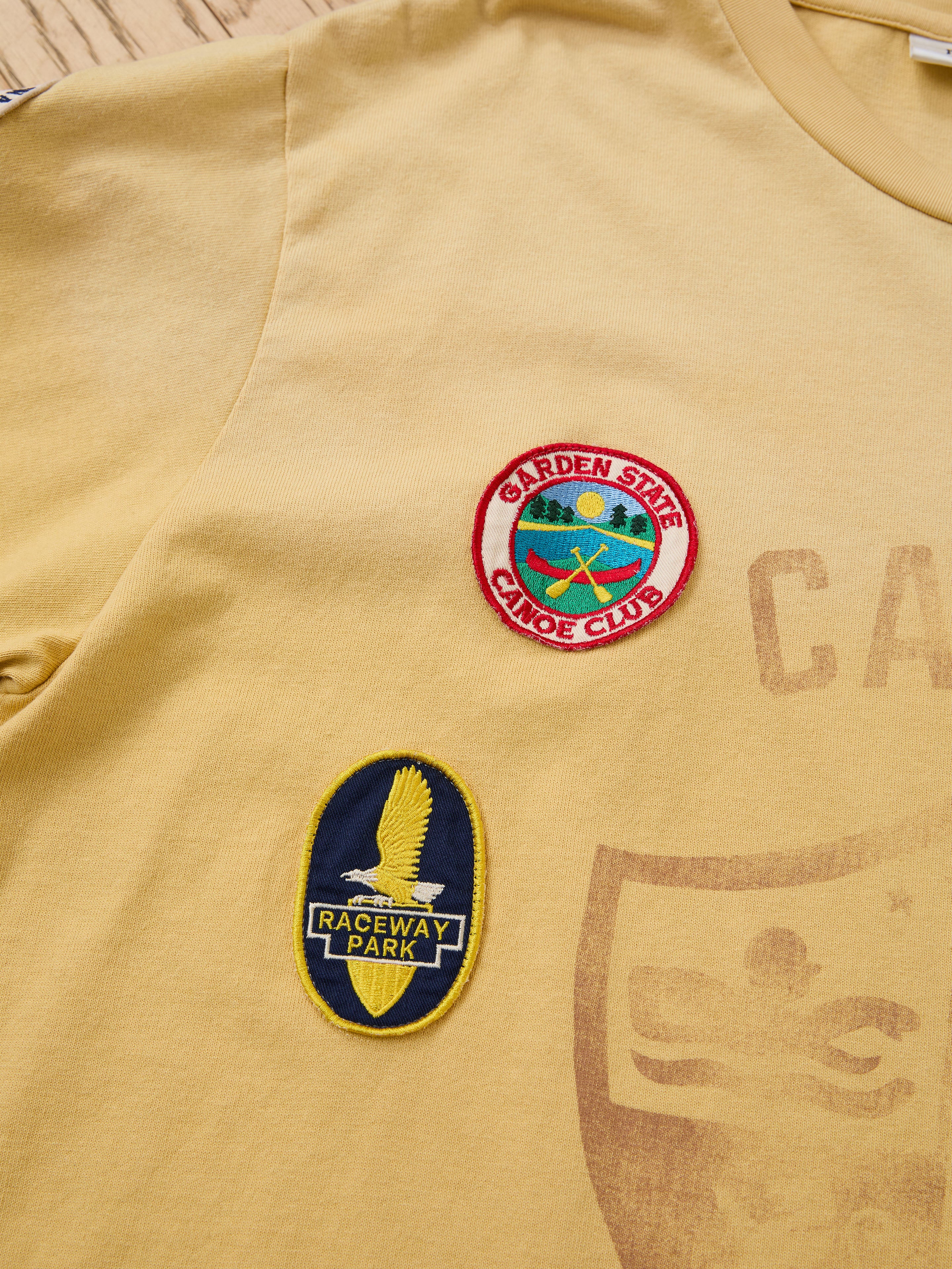 The FOUND Patchwork Camp Tee is a yellow unisex relaxed-fit tee featuring vintage-style embroidered patches: a round "Garden State Canoe Club" and an oval "Raceway Park" patch with an eagle graphic.
