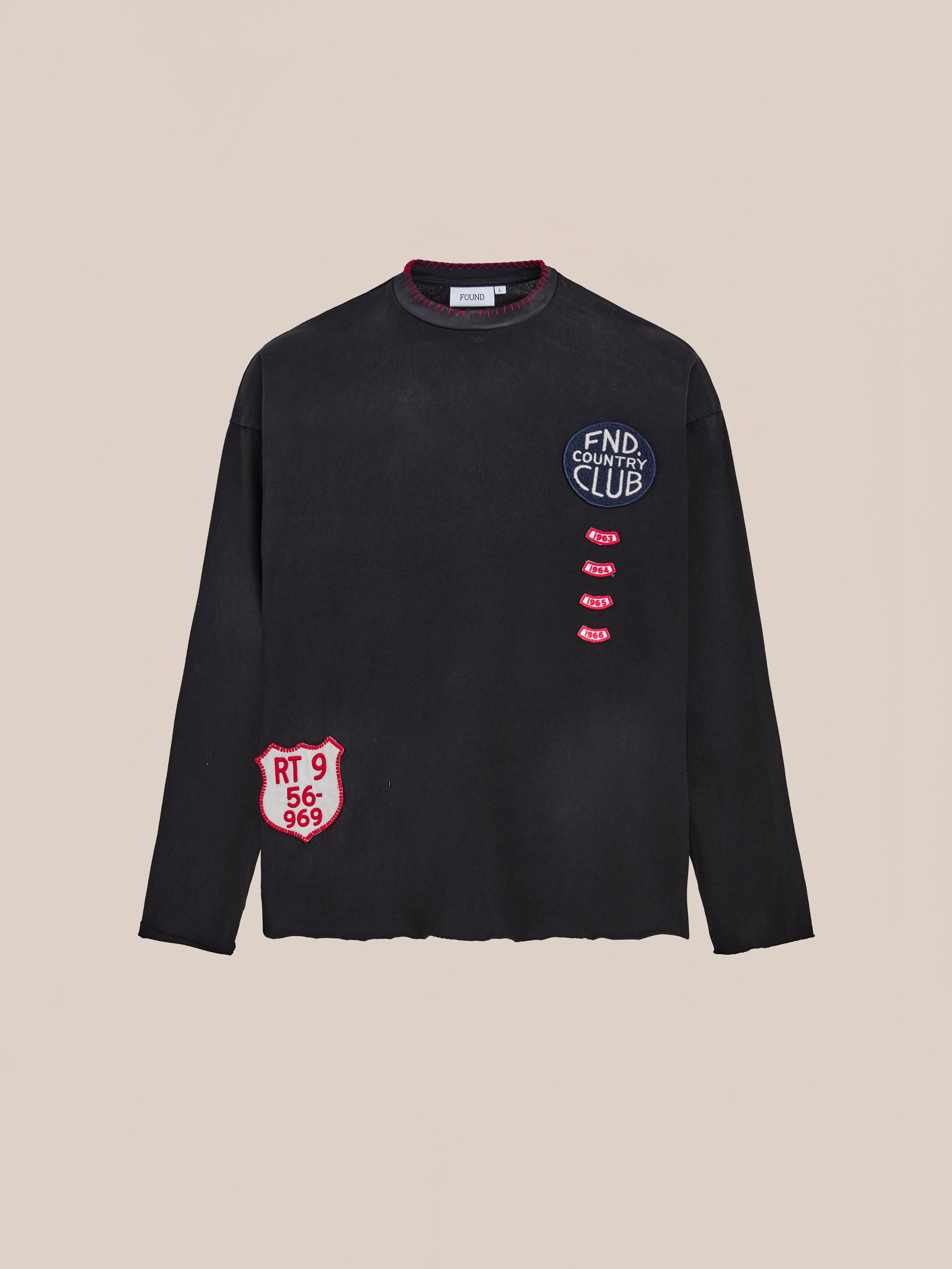 The FOUND Embroidered Patches LS Tee is a vintage washed black long-sleeve shirt featuring distressed details and red and white embroidered patches, including "FND COUNTRY CLUB" on the chest and "RT 9 56-969" near the hem.