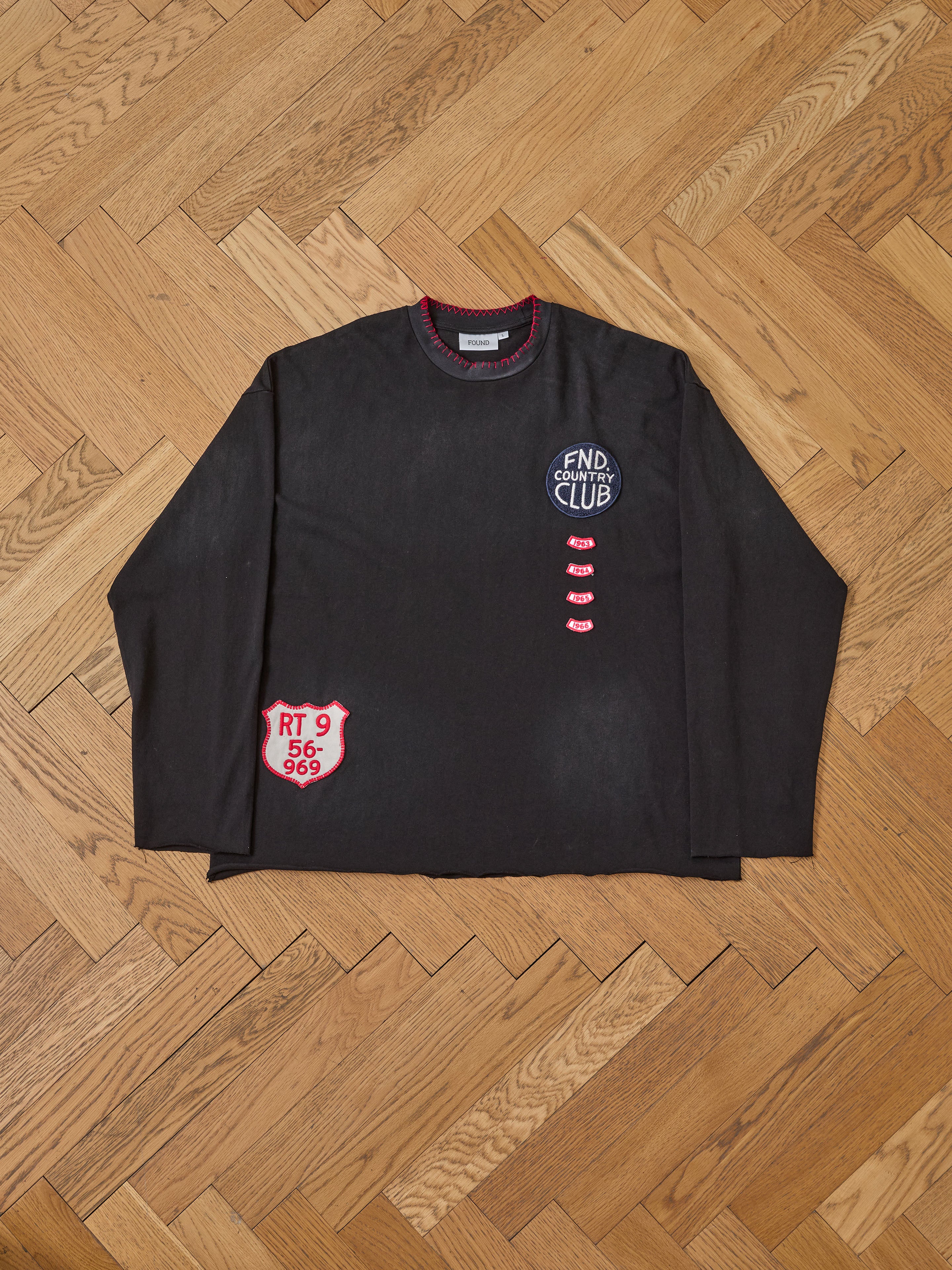 The FOUND Embroidered Patches LS Tee is a vintage washed black long sleeve shirt with red and white embroidered "FND CLUB" and "RT 9 56-969" patches, shown flat on a wooden herringbone floor.