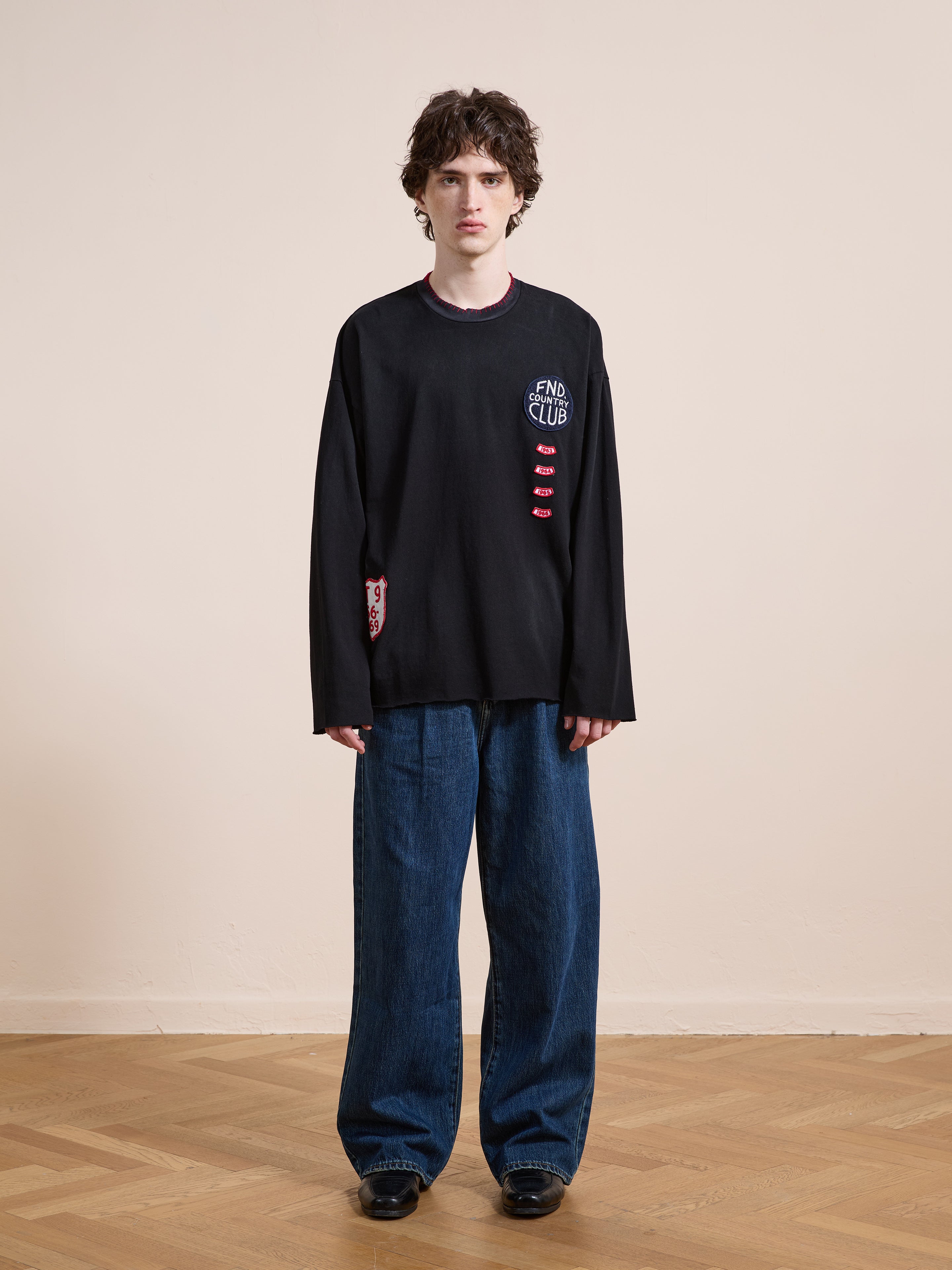 Indoors on a wooden floor, a person wears FOUND’s Embroidered Patches LS Tee in vintage washed black with distressed details and "END CLUB" text, paired with wide blue jeans against a beige wall.
