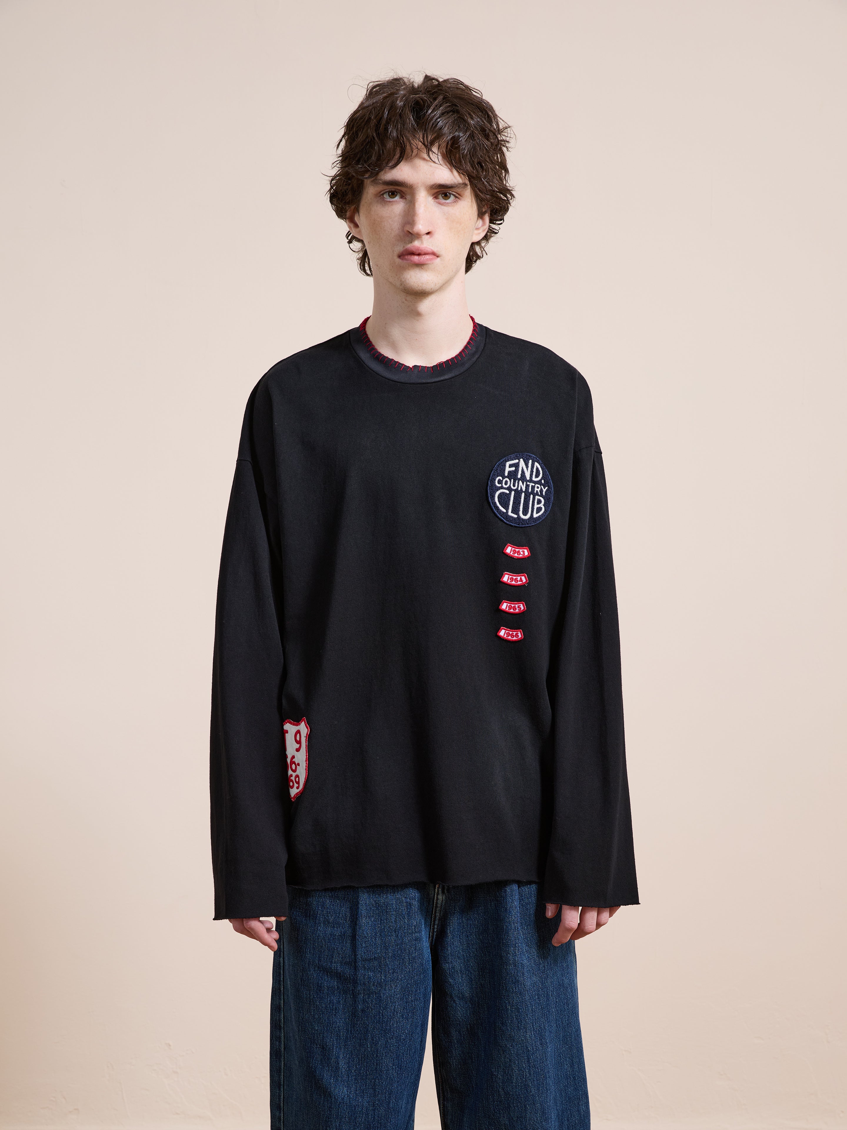 A person with medium-length curly hair wears the FOUND Embroidered Patches LS Tee, a vintage washed black unisex relaxed fit long-sleeve shirt, with blue jeans, standing against a plain beige background.