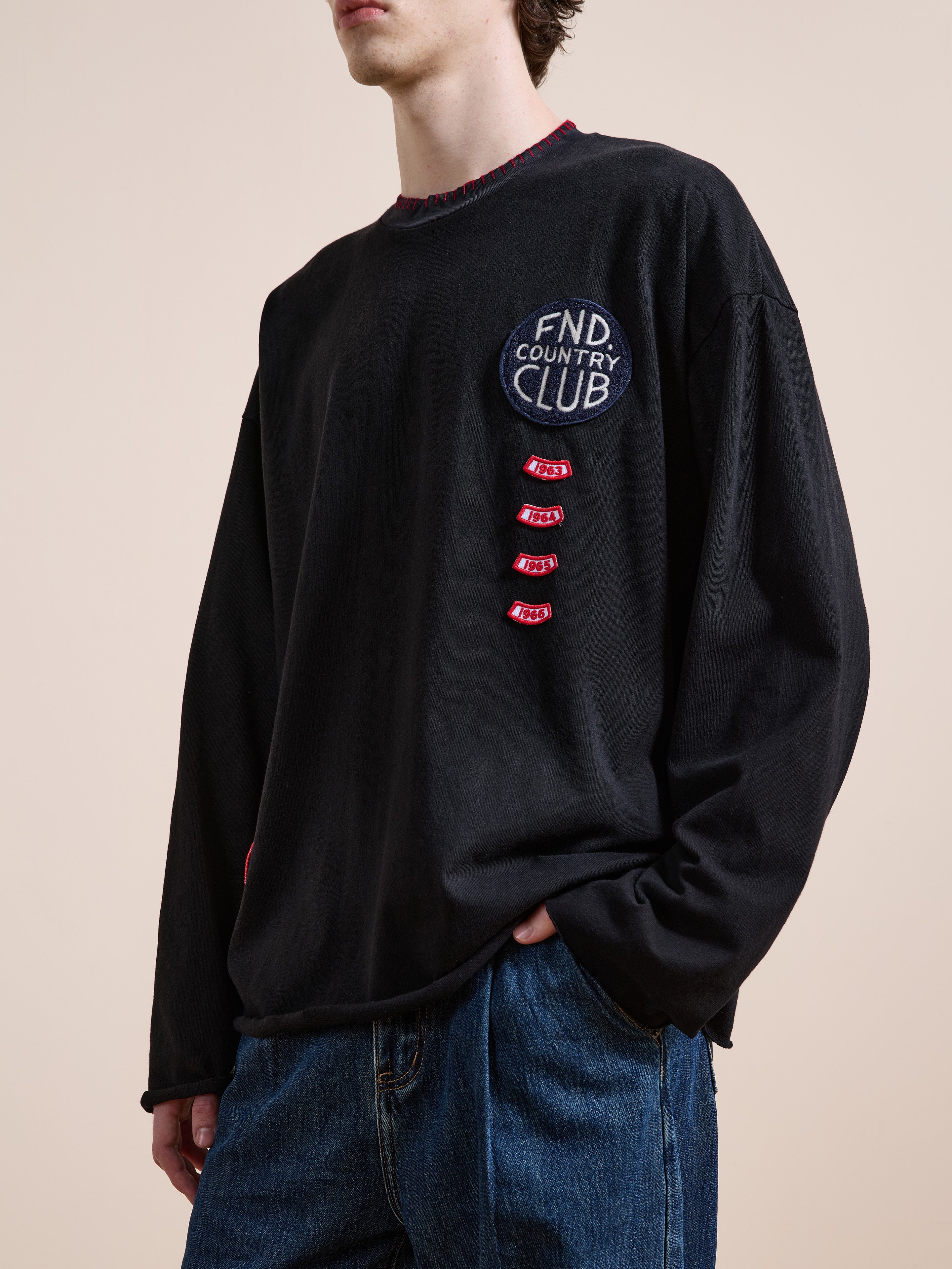 A person wearing the FOUND Embroidered Patches LS Tee in vintage washed black with "FND COUNTRY CLUB" and embroidered patches, paired with blue jeans, stands against a plain beige background.