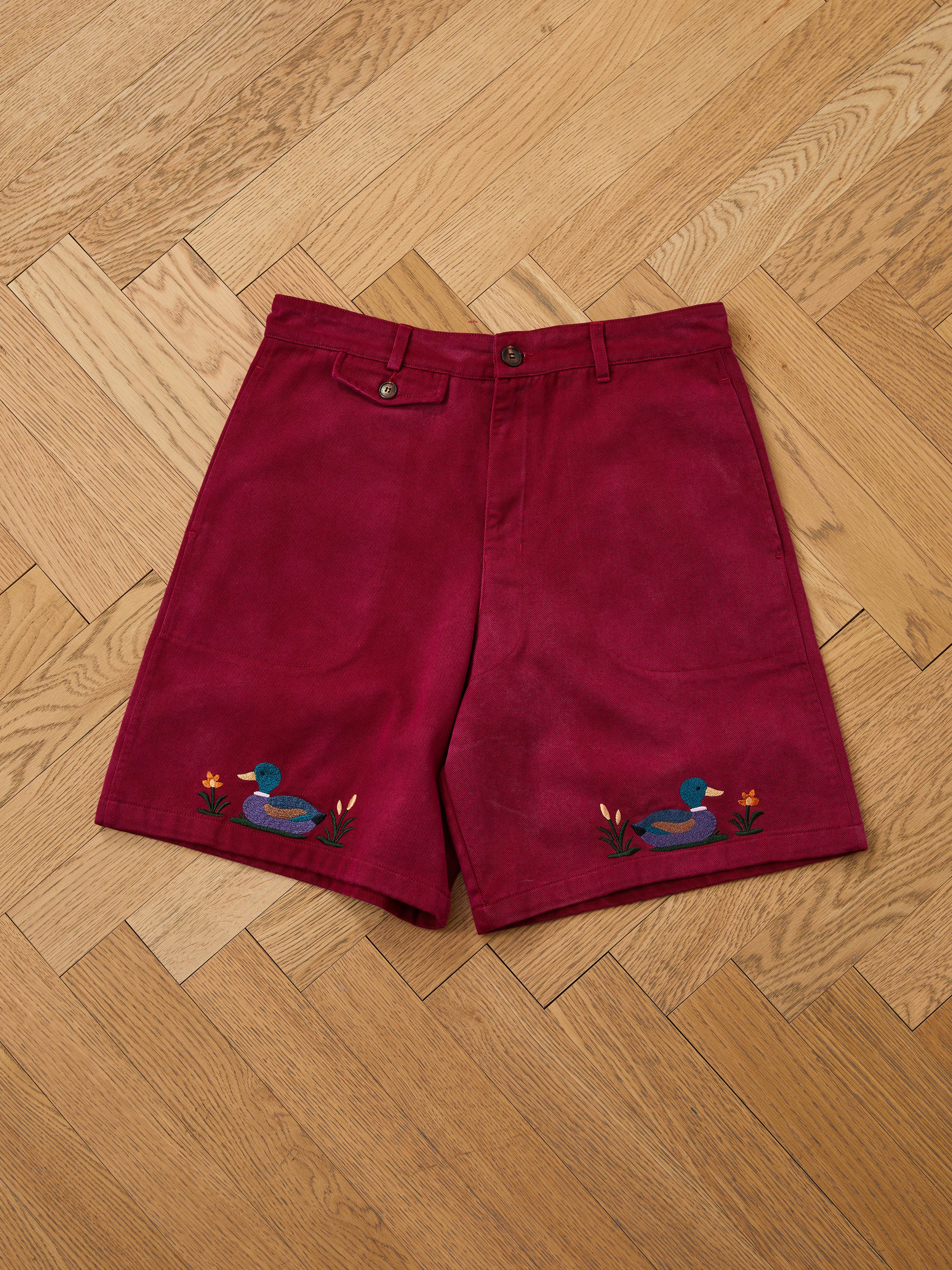 Found Embroidered Duck Shorts: vintage maroon cotton canvas with front button and zipper, featuring blue duck and flower embroidery near the hems, shown laid flat on a wooden herringbone floor.