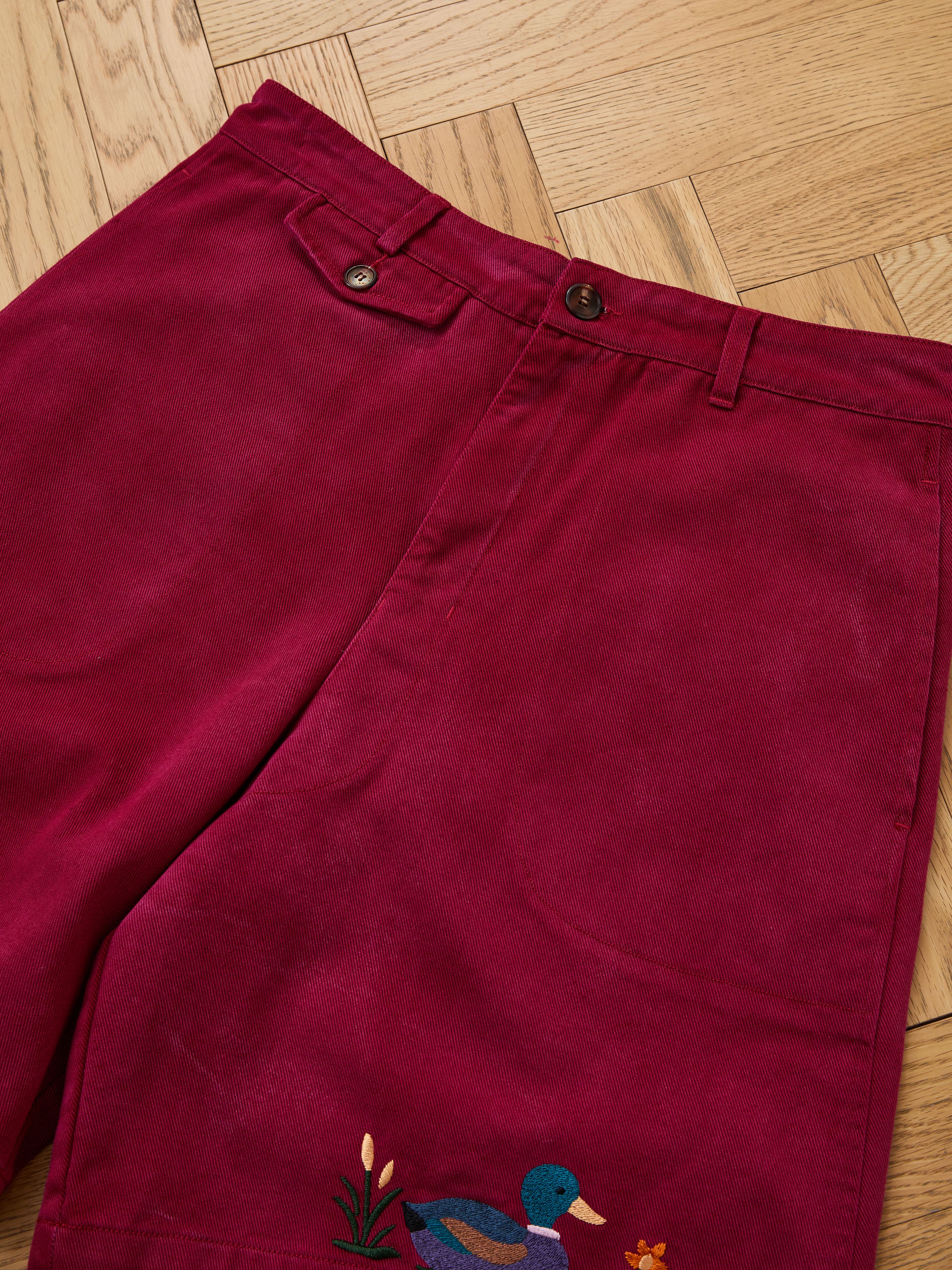 Found Embroidered Duck Shorts in vintage maroon, featuring brown buttons, belt loops, and an embroidered duck with flowers near the pocket, styled flat on a wooden floor for a classic look.