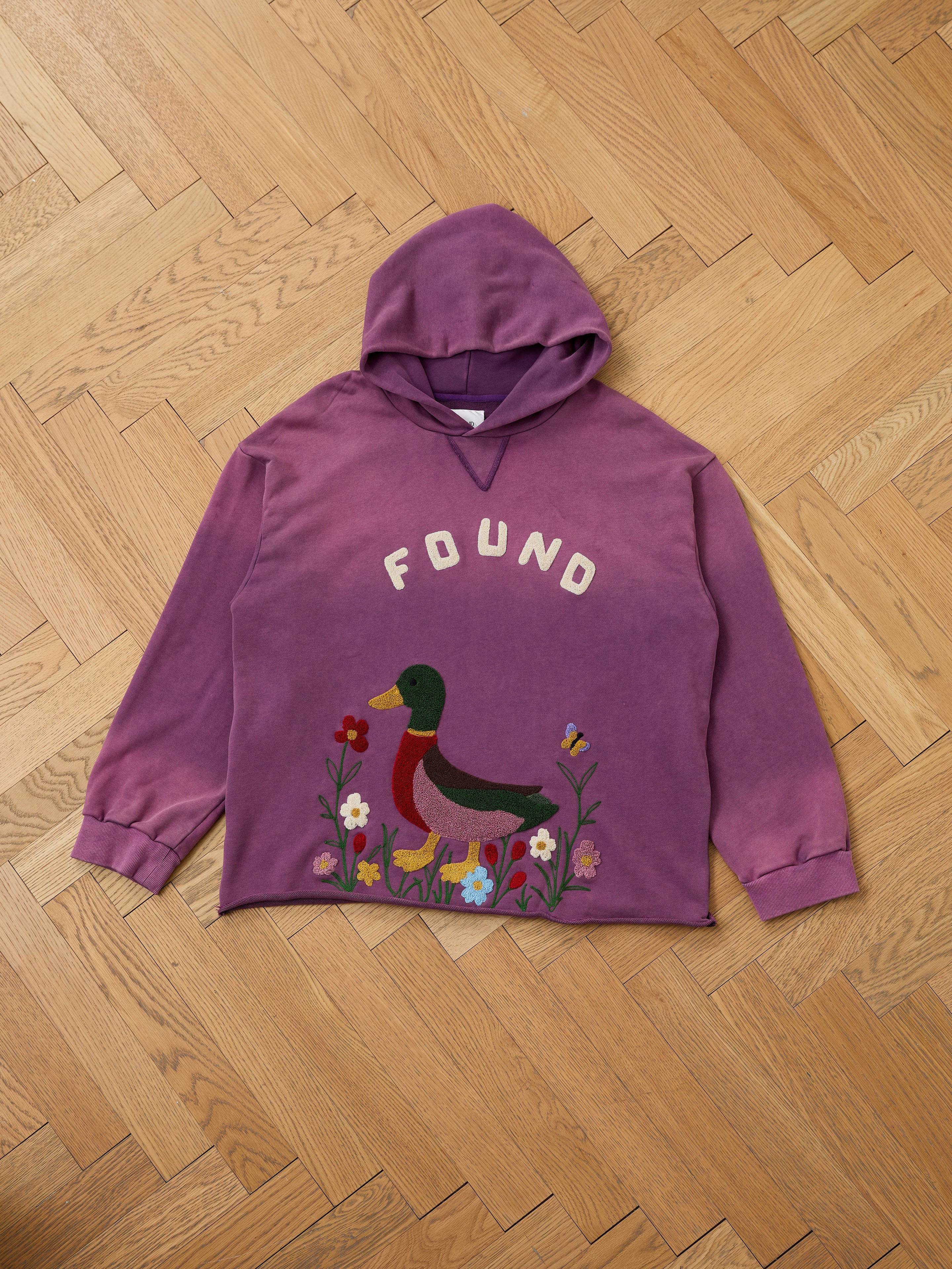 The FOUND Embroidered Wildlife Hoodie features a duck and floral design, shown flat on a herringbone floor. This vintage French-terry hoodie is perfect for wildlife lovers seeking comfort and unique style.