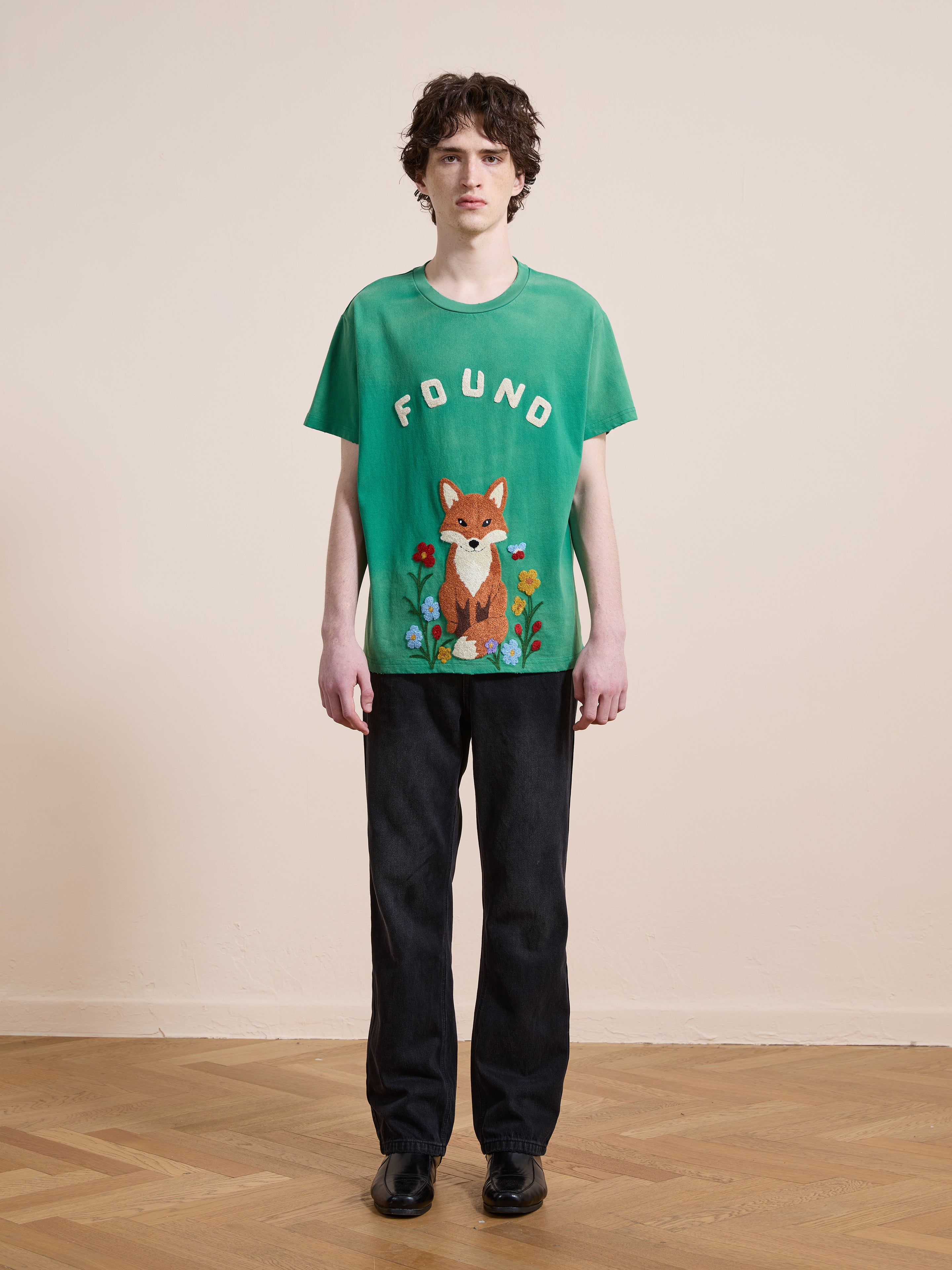 A person stands indoors against a plain wall, wearing a FOUND Embroidered Wildlife Tee paired with black pants and black shoes.