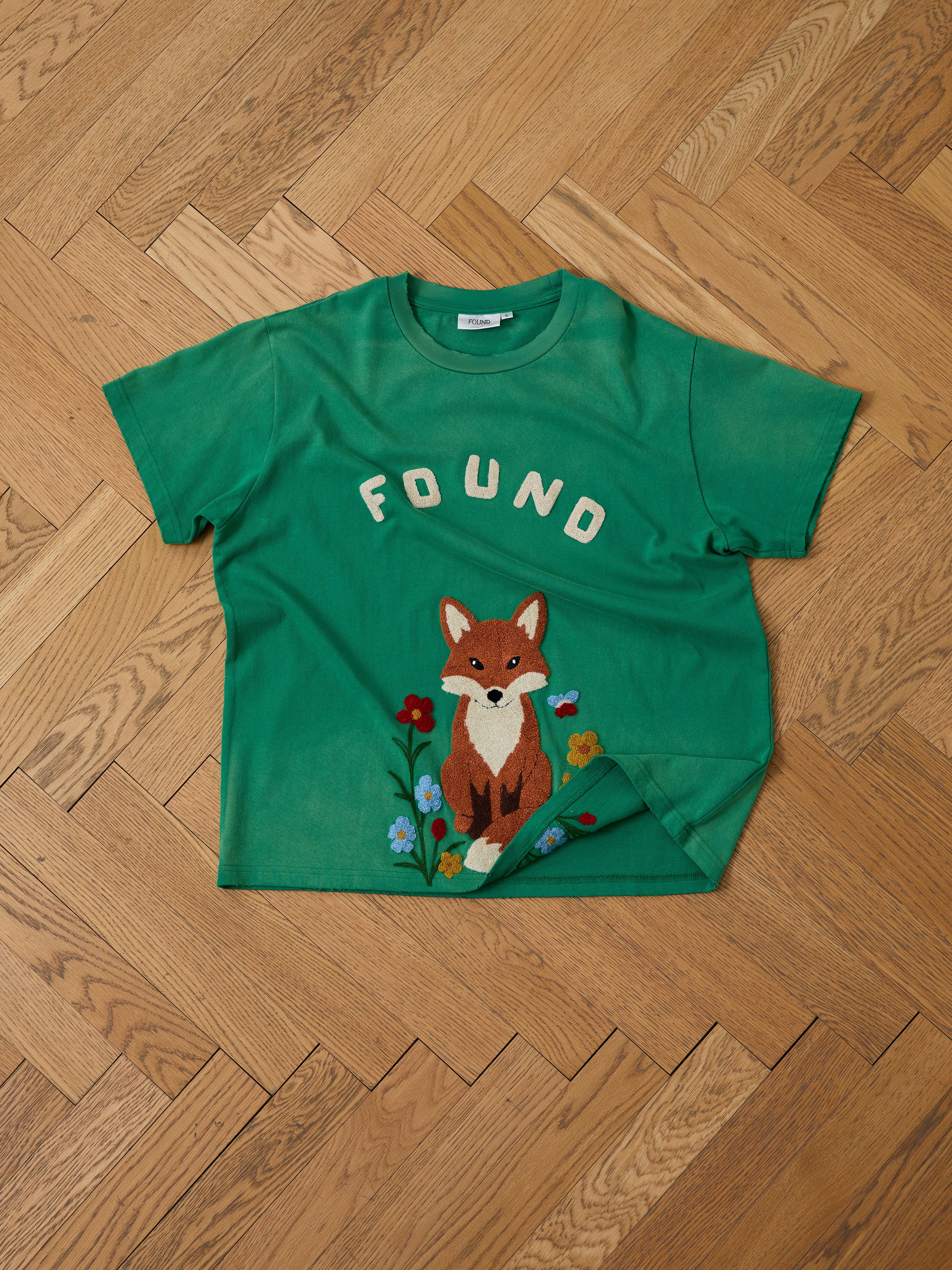 The FOUND Embroidered Wildlife Tee features a green vintage wash, a countryside fox among flowers illustration, and "FOUND" embroidery, shown laid flat on a wooden herringbone floor.