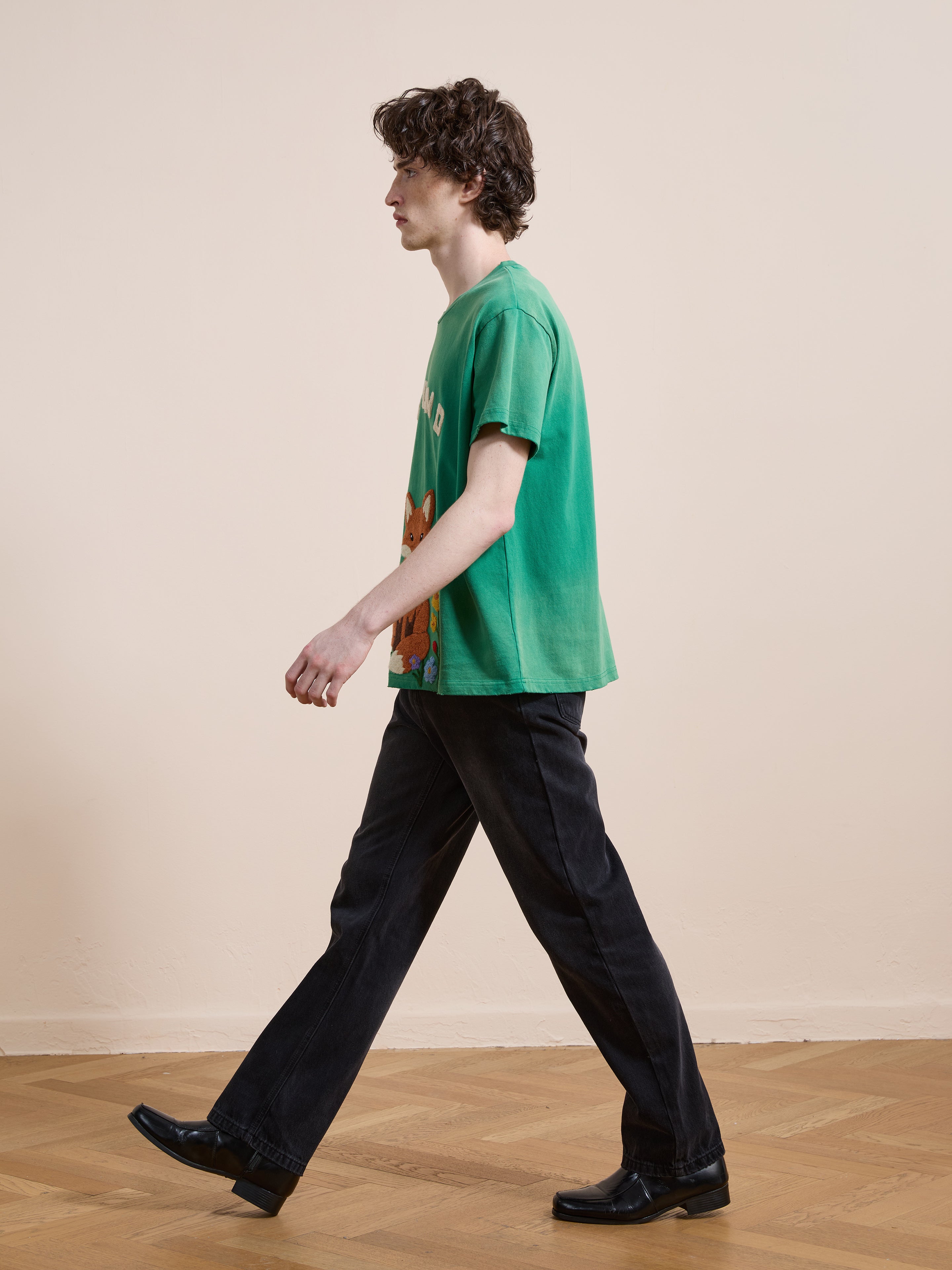 A person with curly hair, wearing the FOUND Embroidered Wildlife Tee in a unisex relaxed fit, black pants, and black shoes, walks indoors on a wooden floor.
