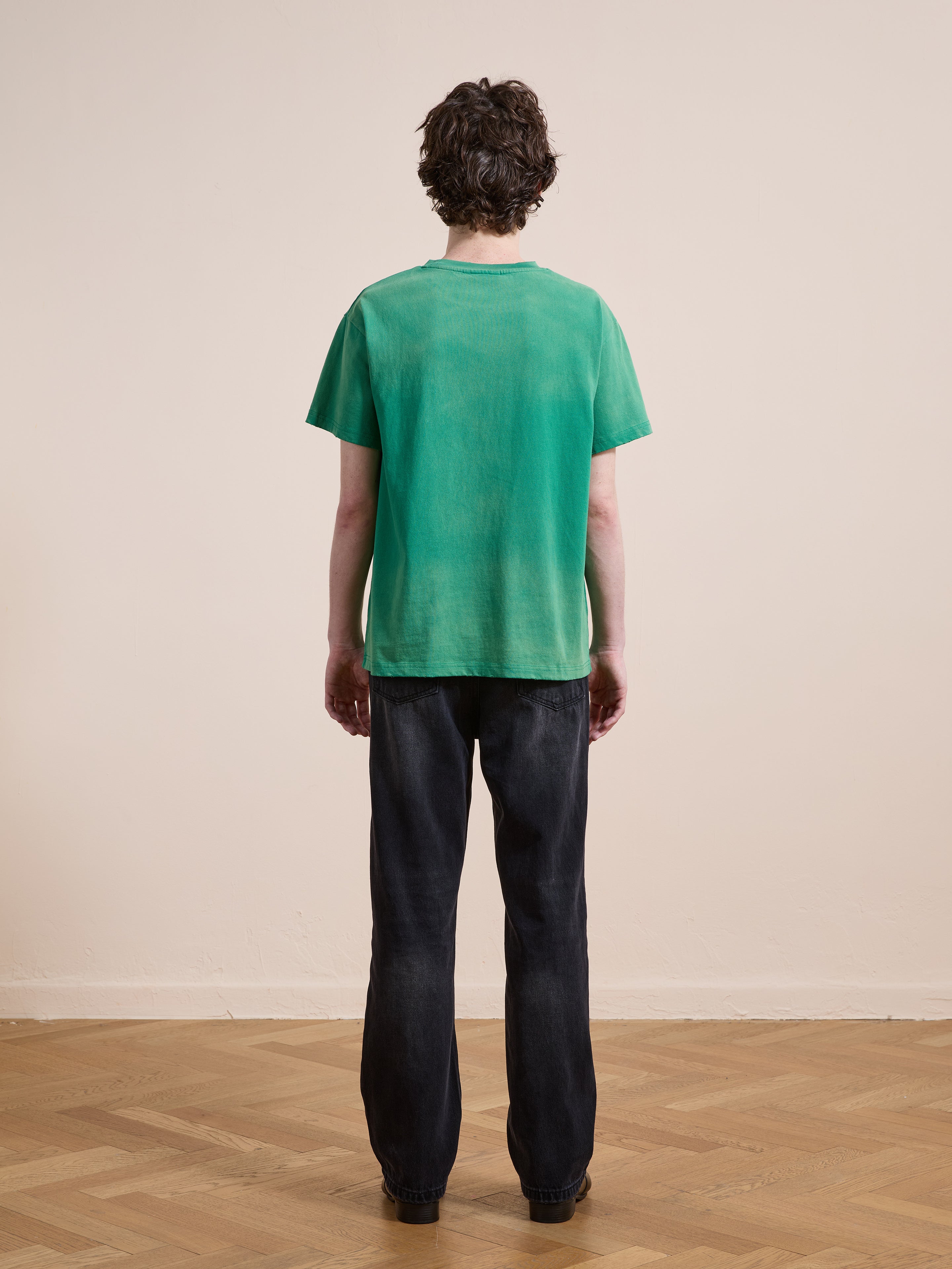 A person with short dark hair stands indoors on a wood floor, facing away, wearing a FOUND Embroidered Wildlife Tee in green and black jeans.