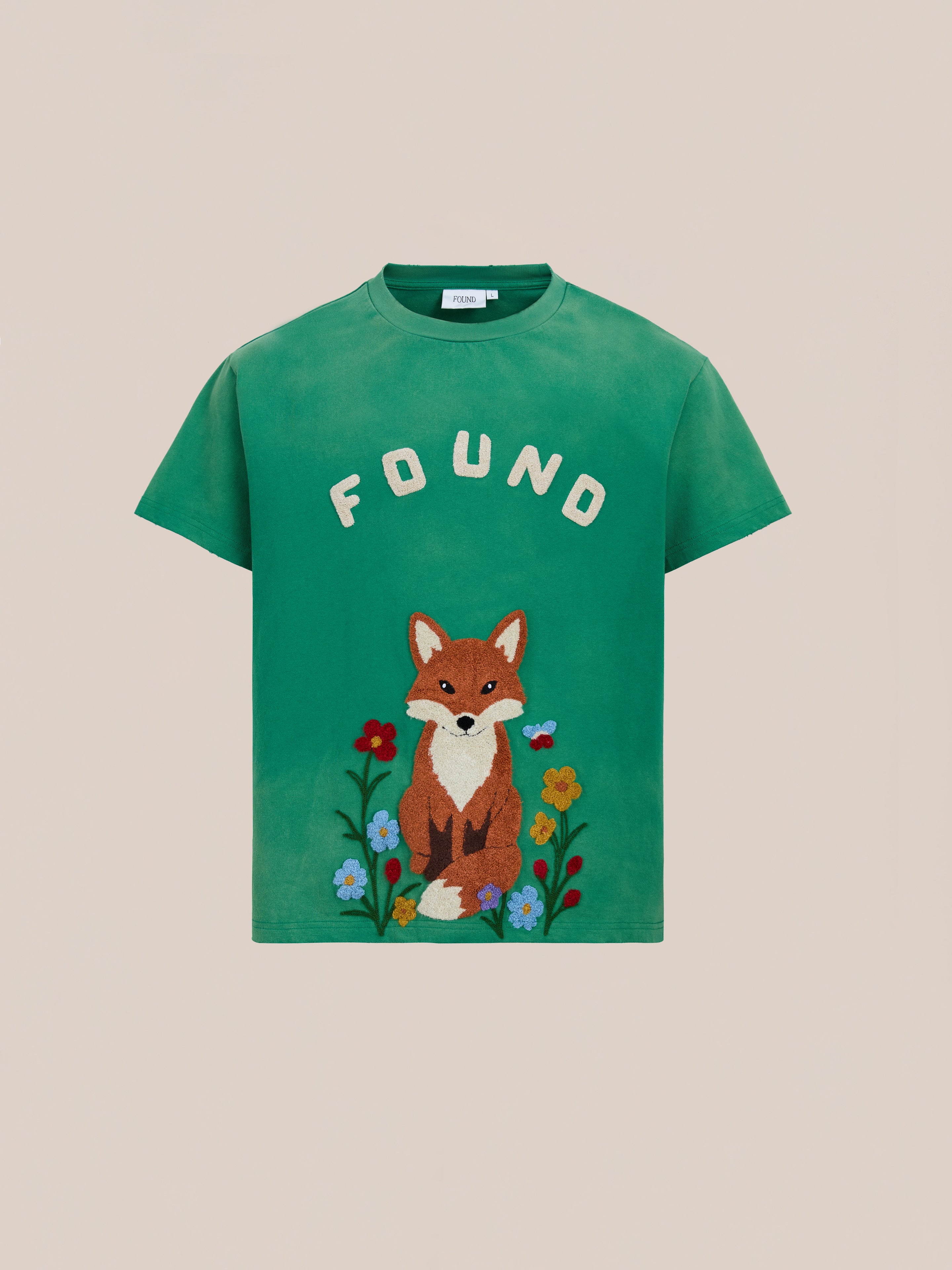 The Embroidered Wildlife Tee by FOUND features a green vintage wash with an embroidered fox motif sitting among flowers and the "FOUND" logo above.