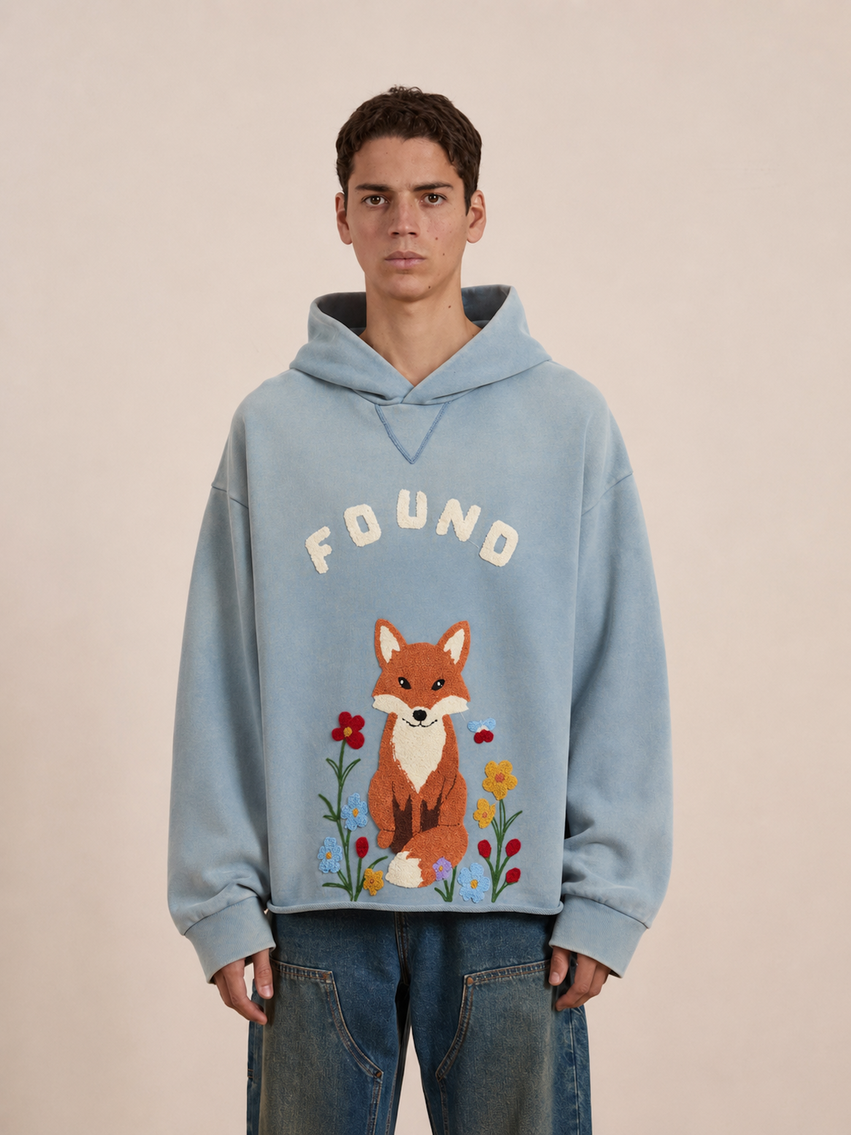 A person wearing a light blue Embroidered Wildlife Hoodie from FOUND, featuring the word “FOUND” and an embroidered fox with flowers, standing against a plain background.