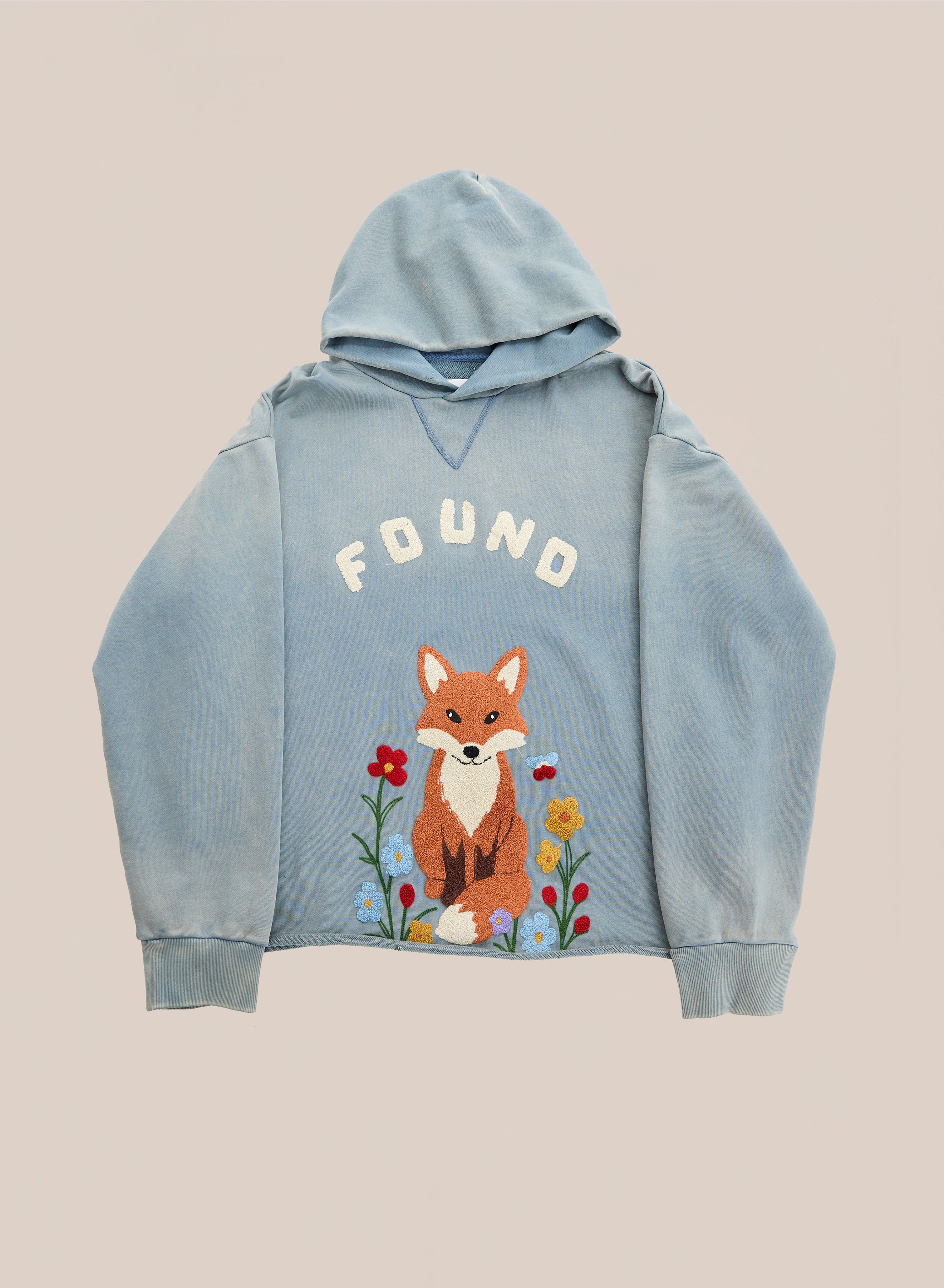 The Embroidered Wildlife Hoodie by FOUND is a light blue vintage French-terry hoodie featuring the brand’s “FOUND” text above a beautifully embroidered fox graphic sitting among flowers, shown on a plain background.