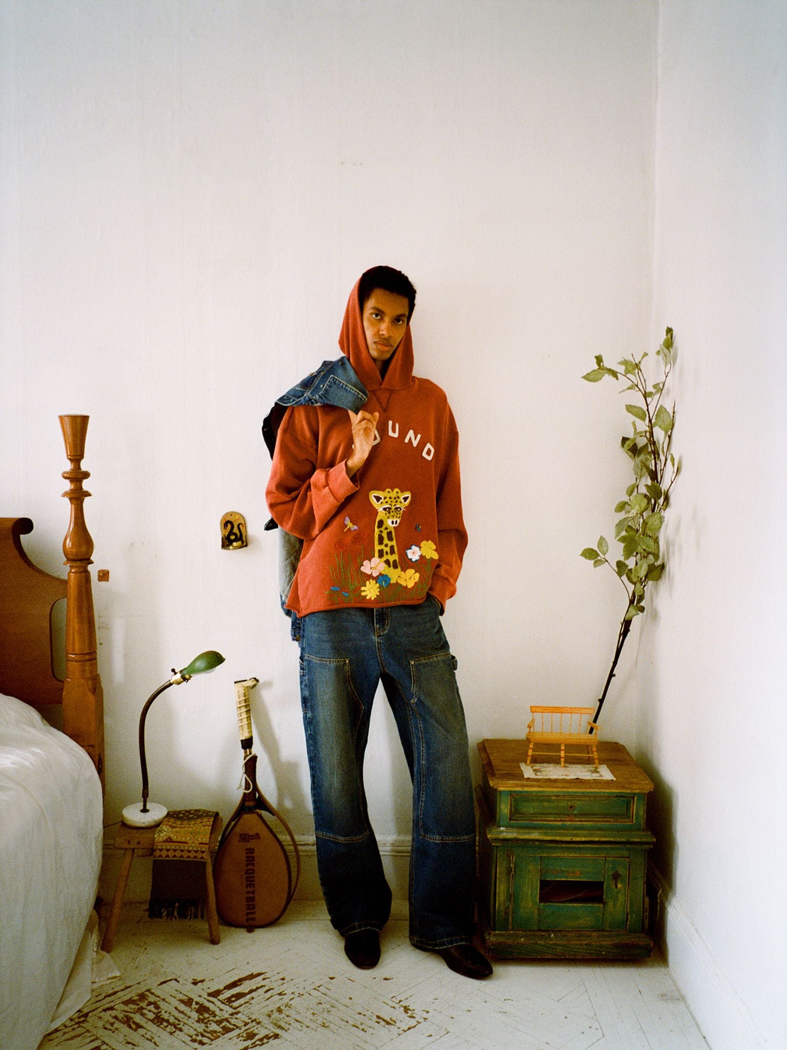 A person in a relaxed fit red FOUND Embroidered Wildlife Hoodie featuring a giraffe graphic stands in a bedroom, holding a denim jacket over their shoulder beside a green nightstand and tennis racket.