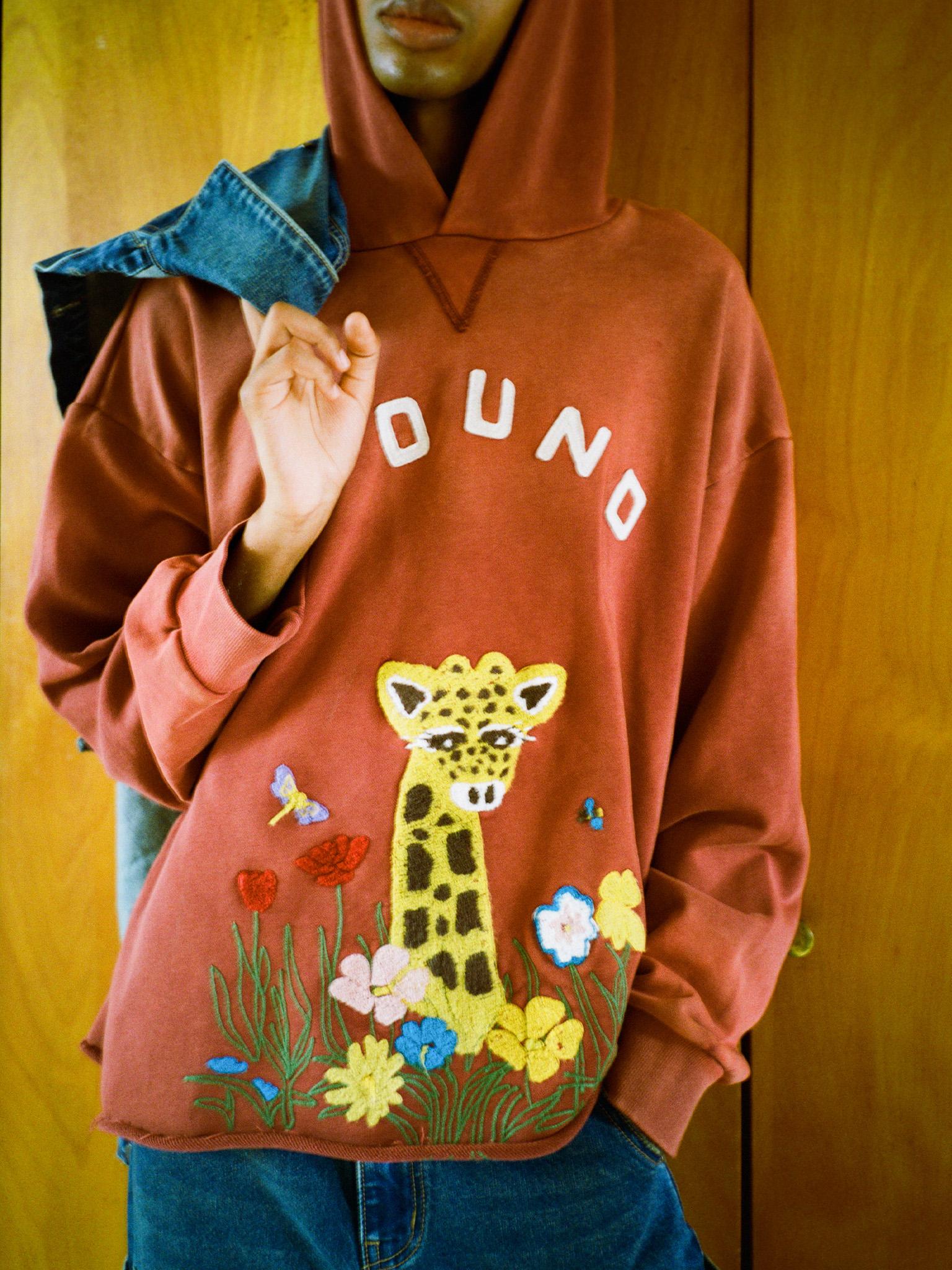 A person wears a relaxed fit, rust Embroidered Wildlife Hoodie by FOUND with "FOUND" text, wildlife chenille embroidery, floral accents, and a giraffe design, holding a denim jacket over their shoulder against a wooden backdrop.