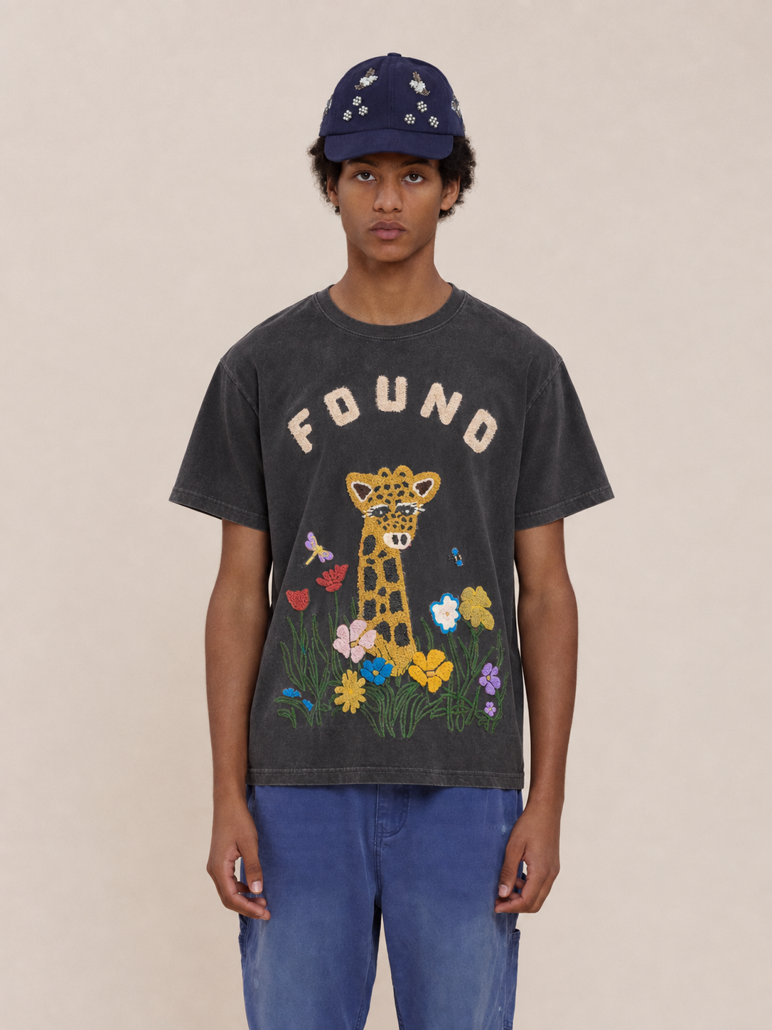 Young person in a navy cap and Embroidered Wildlife Tee by FOUND, featuring flowers, butterflies, and “FOUND,” standing against a beige background.