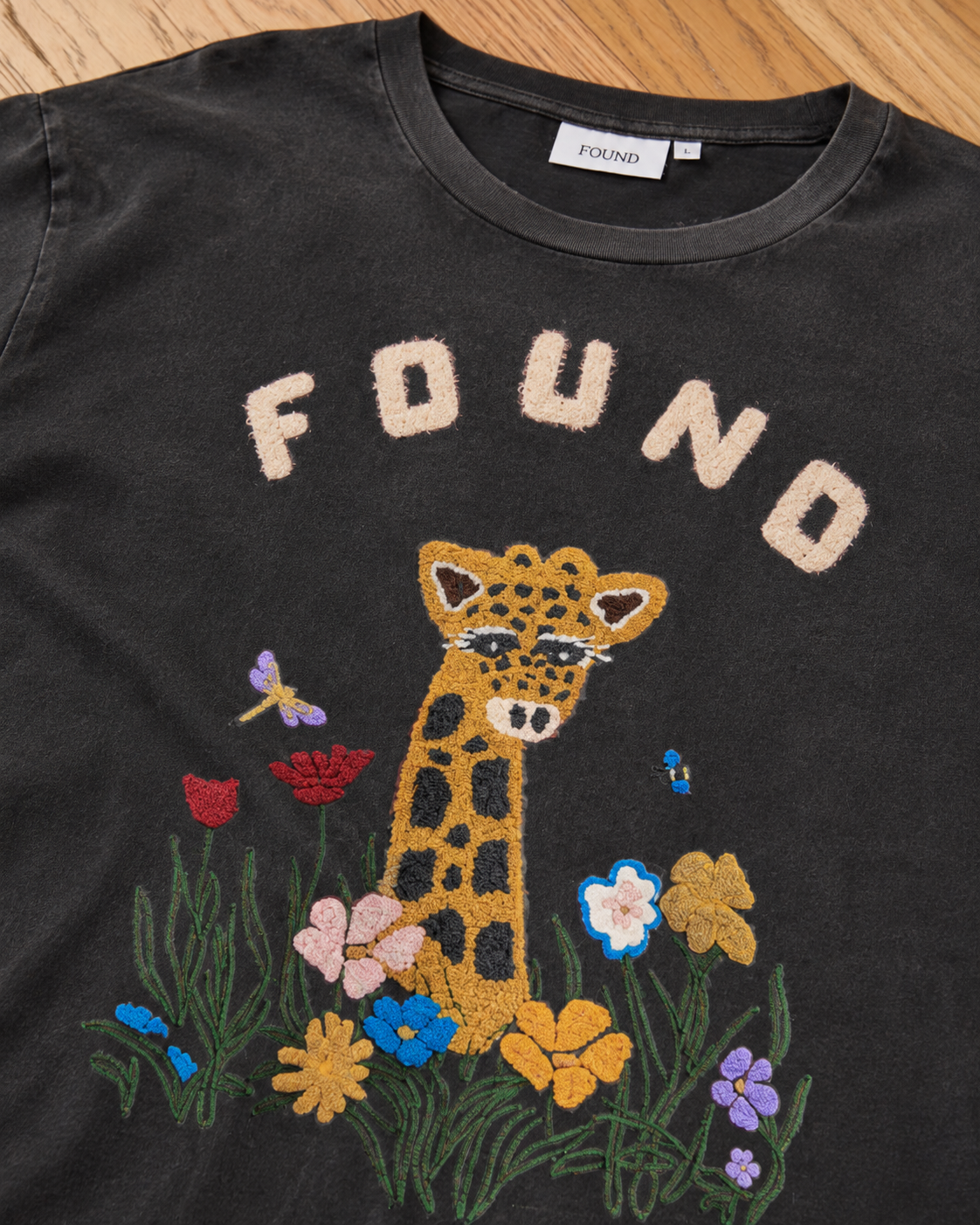 The Embroidered Wildlife Tee by FOUND features “FOUND” text, chenille embroidery of a giraffe, colorful flowers, a butterfly, and a bee on the front.