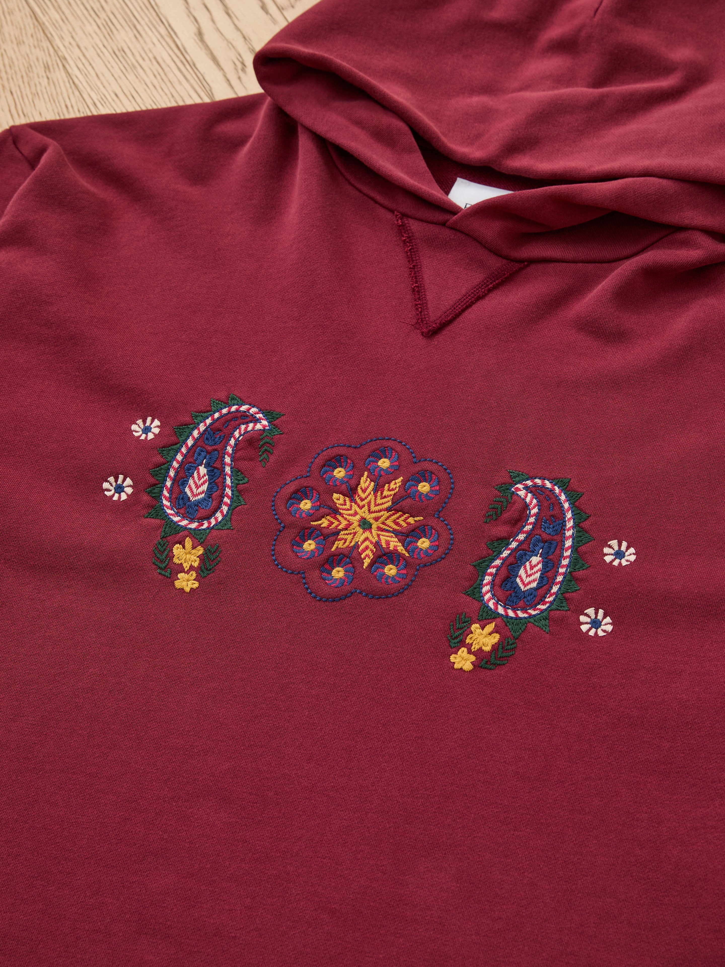 The FOUND Embroidered Kantha Paisley Hoodie is a maroon unisex cotton fleece with vibrant paisley and floral embroidery on the front, shown laid flat on a wooden surface.